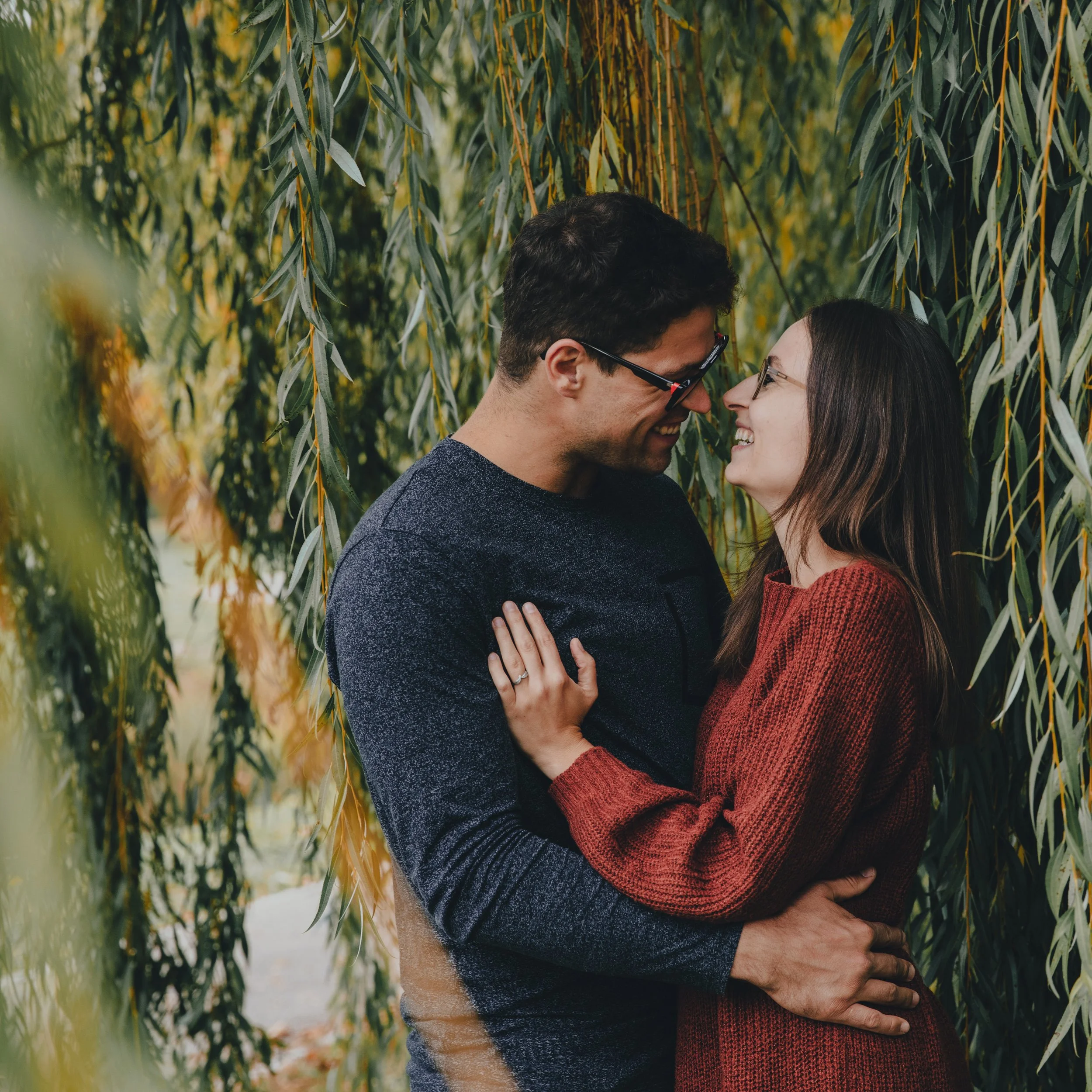 Ein Paar umarmt sich, lächelt und schaut sich liebevoll in die Augen, umgeben von grünen Blättern im Freien.