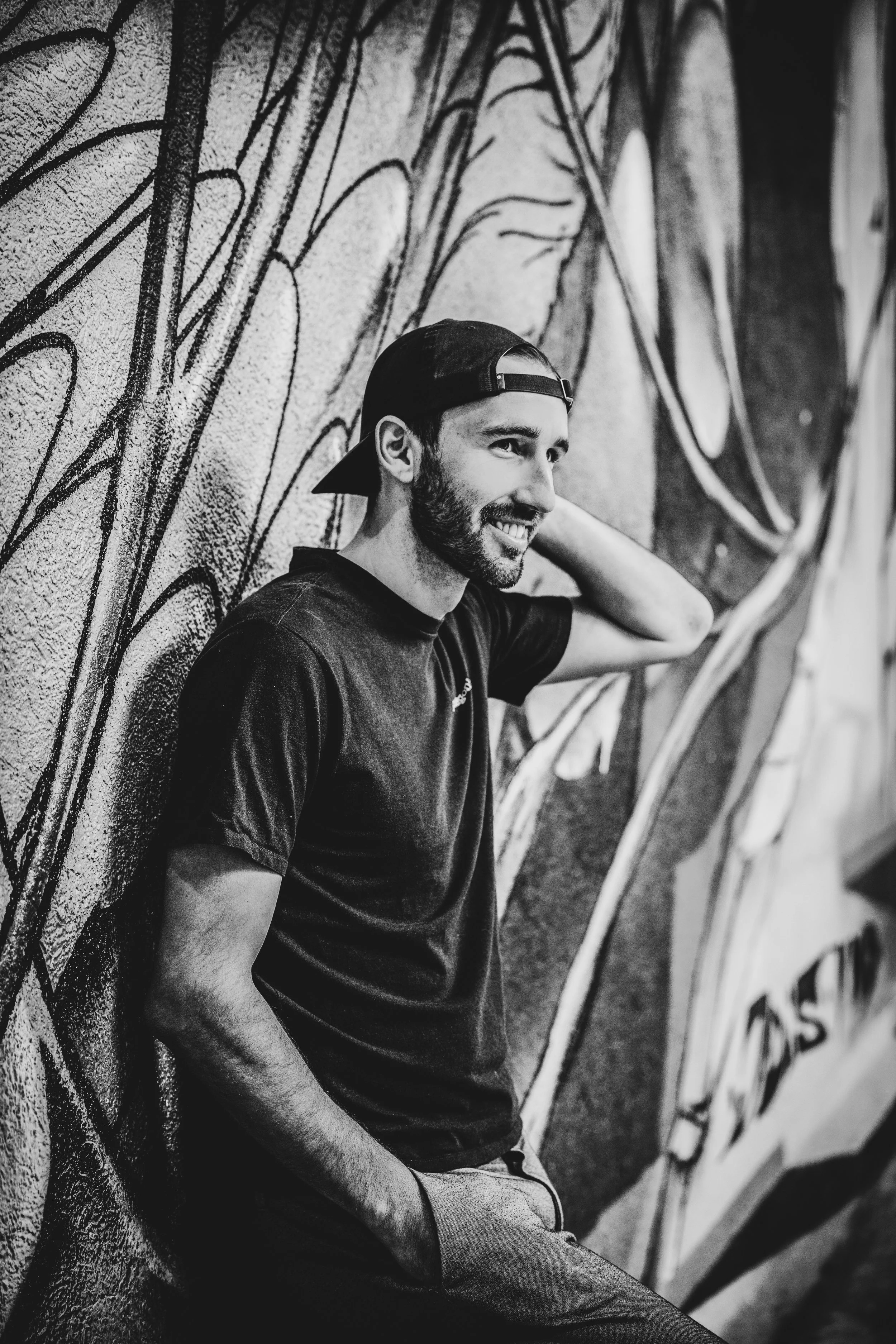 Ein lächelnder junger Mann mit Bart, schwarzer Hautschutzkappe, schwarzem T-Shirt und grauen Hosen lehnt an einer Wand mit Graffiti.