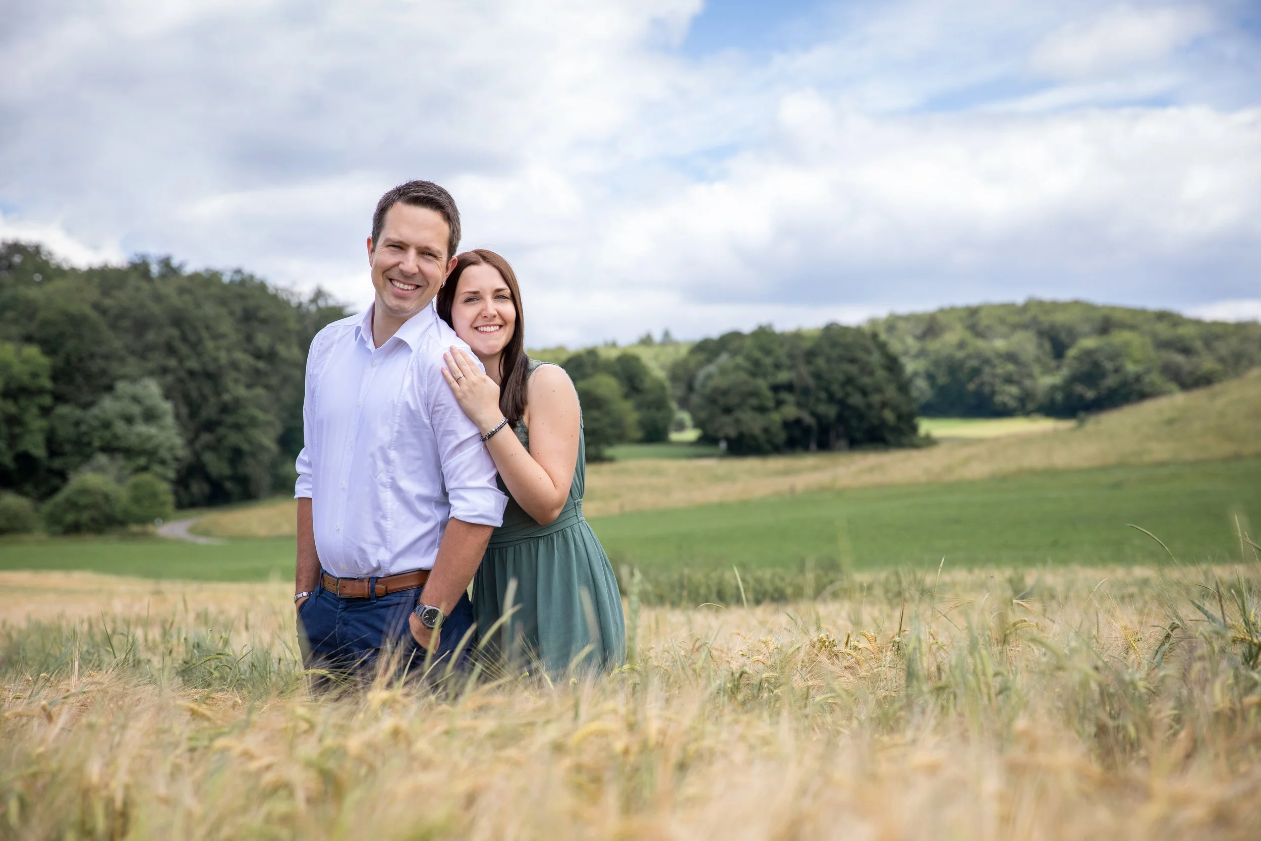 Ein lachendes Paar steht in einem goldgelben Weizenfeld, im Hintergrund grüne Bäume und ein blauer Himmel mit Wolken.