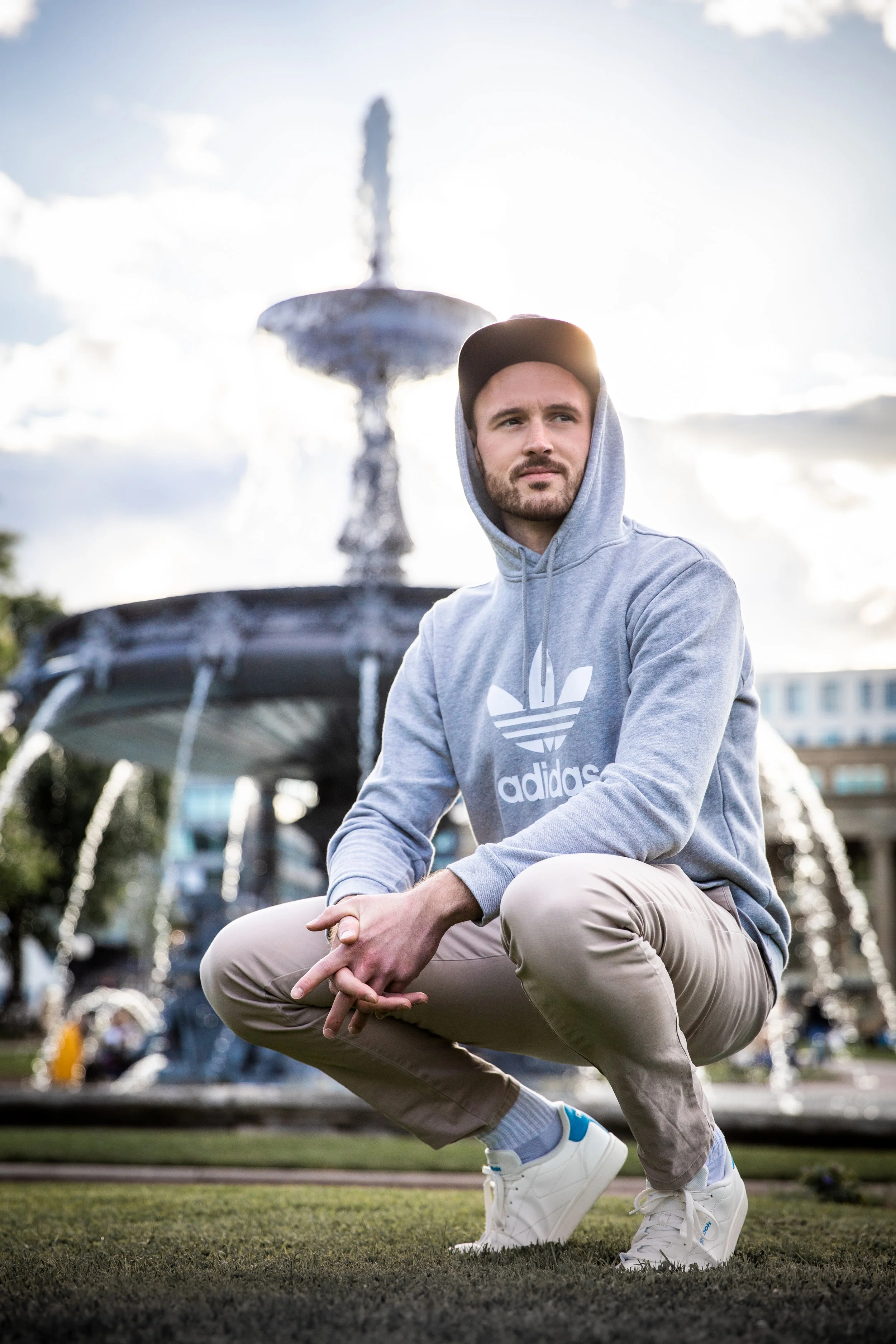 Junger Mann in grauem Adidas-Hoodie und beige Hosen sitzt vor einem Springbrunnen im Freien, bei bewölktem Himmel.