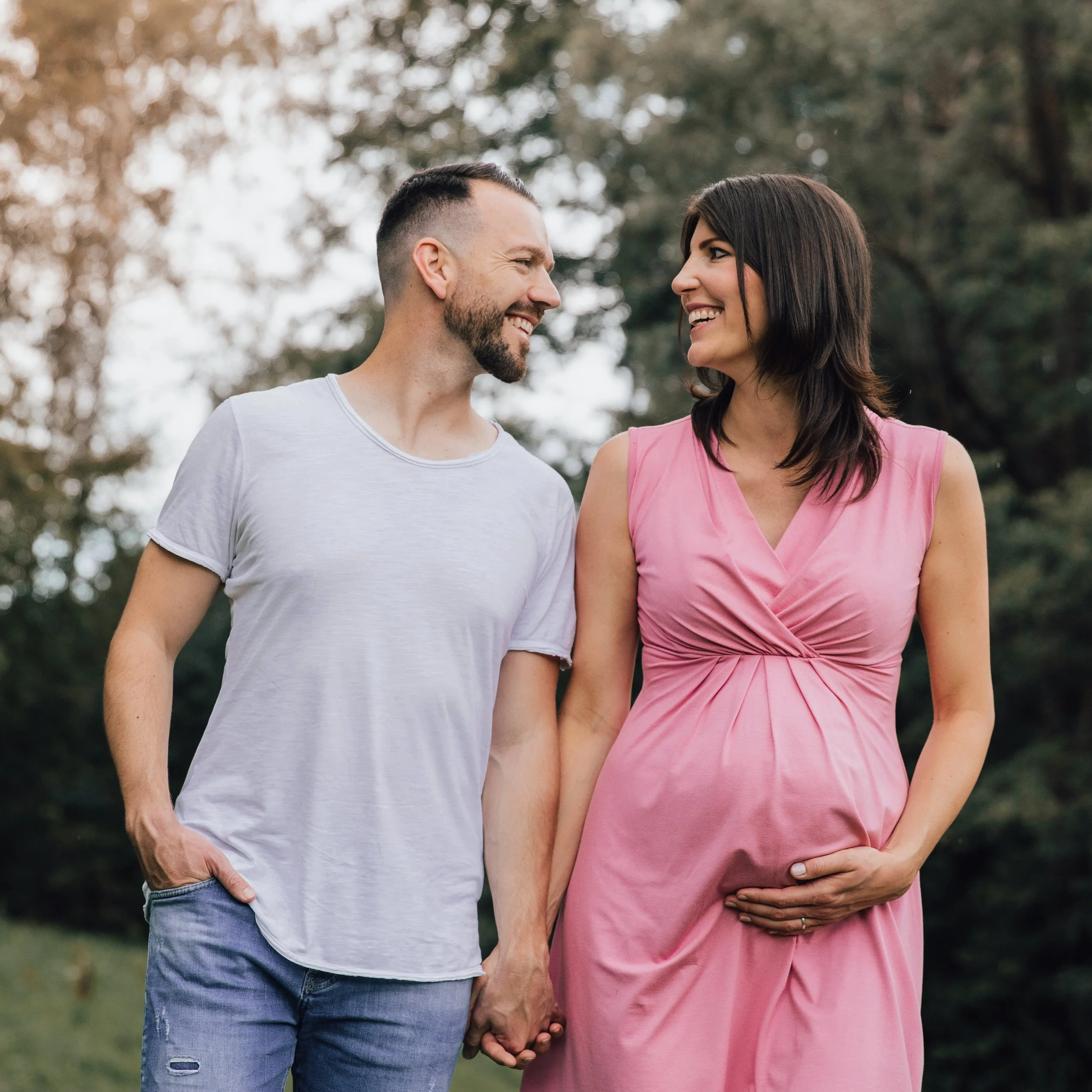 Ein glückliches Paar hält Händchen im Park, die schwangere Frau trägt ein pinkes Kleid, der Mann trägt ein weißes T-Shirt.