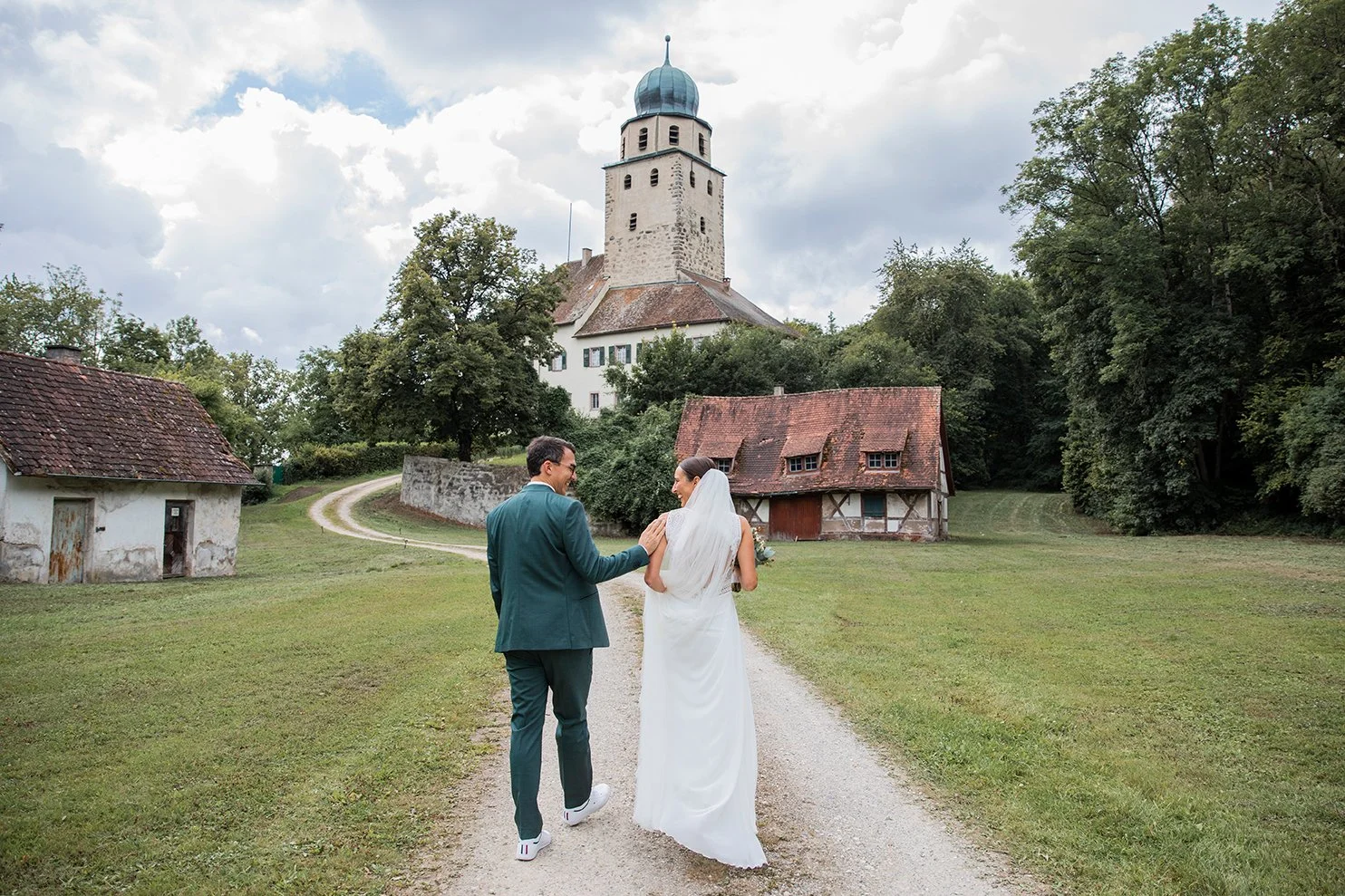Ein frisch verheiratetes Paar, der Bräutigam und die Braut, laufen eine unbefestigte Straße entlang, umgeben von grünen Wiesen und alten Gebäuden, mit einer mittelalterlichen Kirche im Hintergrund, unter einem bewölkten Himmel.