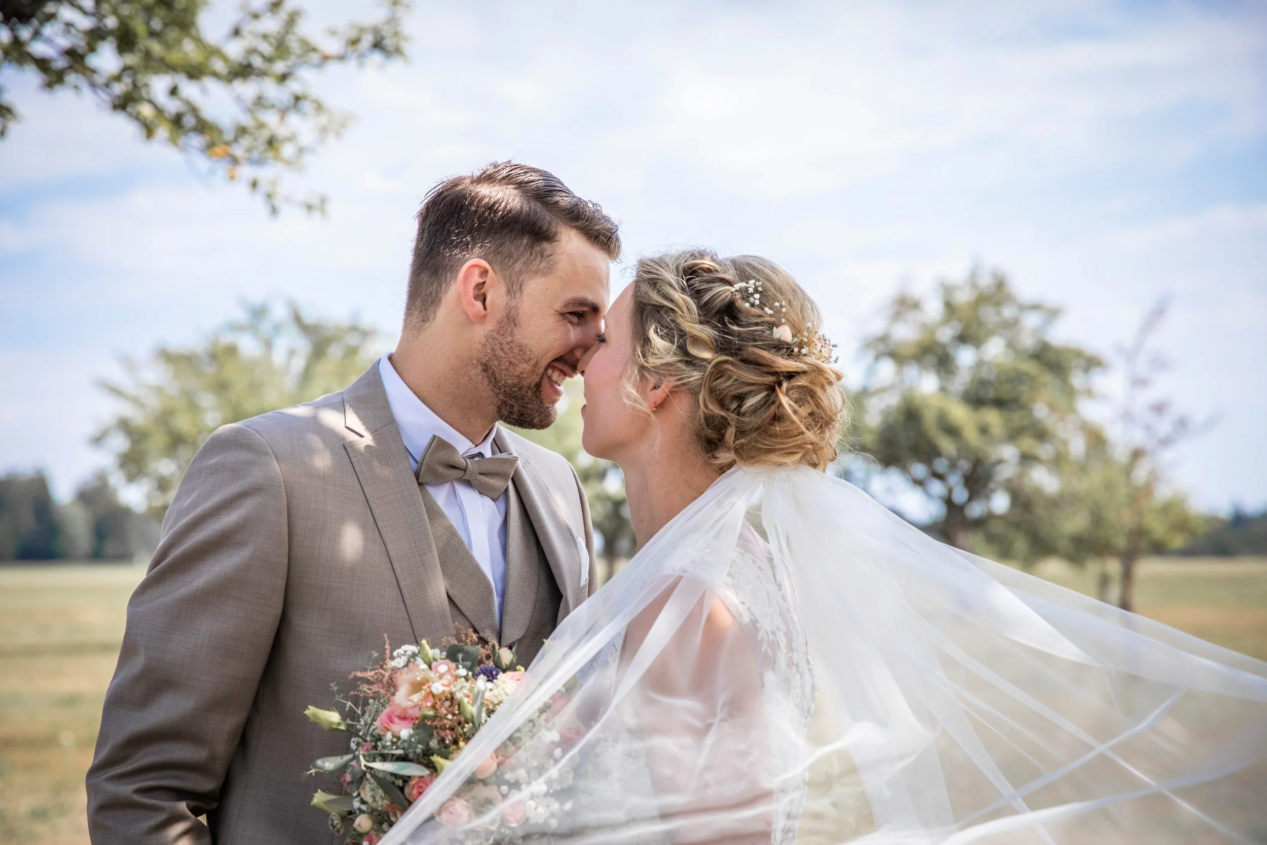 Ein Brautpaar auf einem Feld bei Sonnenschein, die Braut hält einen Blumenstrauß.