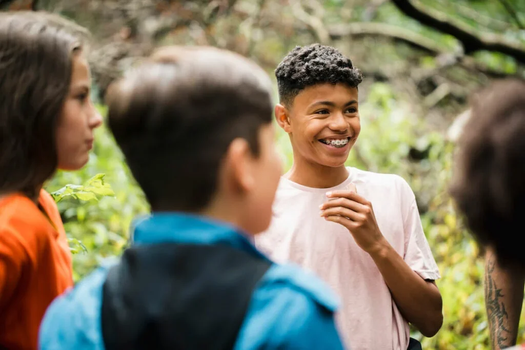 A group of teenagers talking outdoors in a wooded area, with one boy smiling and holding his hand to his chest.