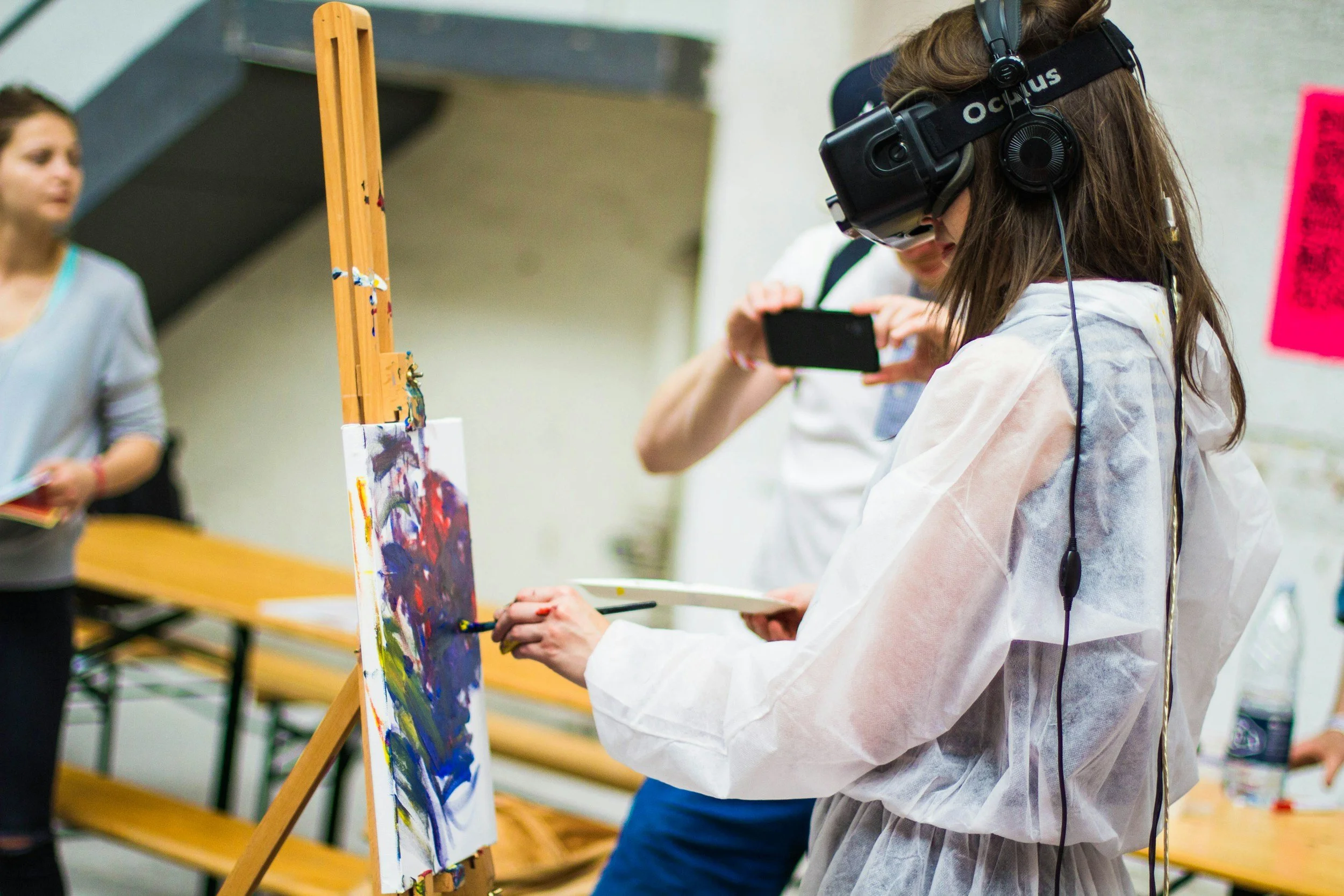 A woman wearing VR goggles painting on a canvas with a paintbrush, with people around taking photos and holding books in an indoor setting.