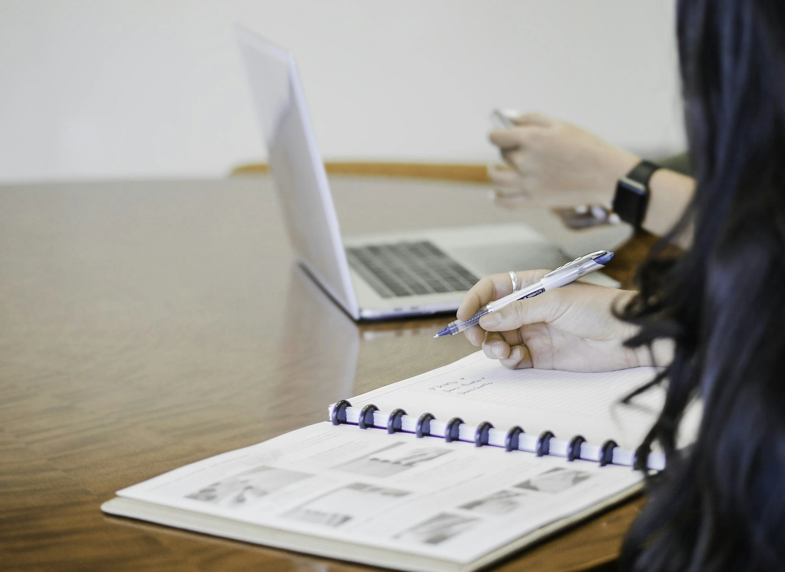 Person taking notes with a pen, open comic book and laptop on a wooden table.