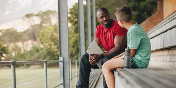 A man and a boy sitting on stadium bleachers, talking, with some trees and a building in the background.