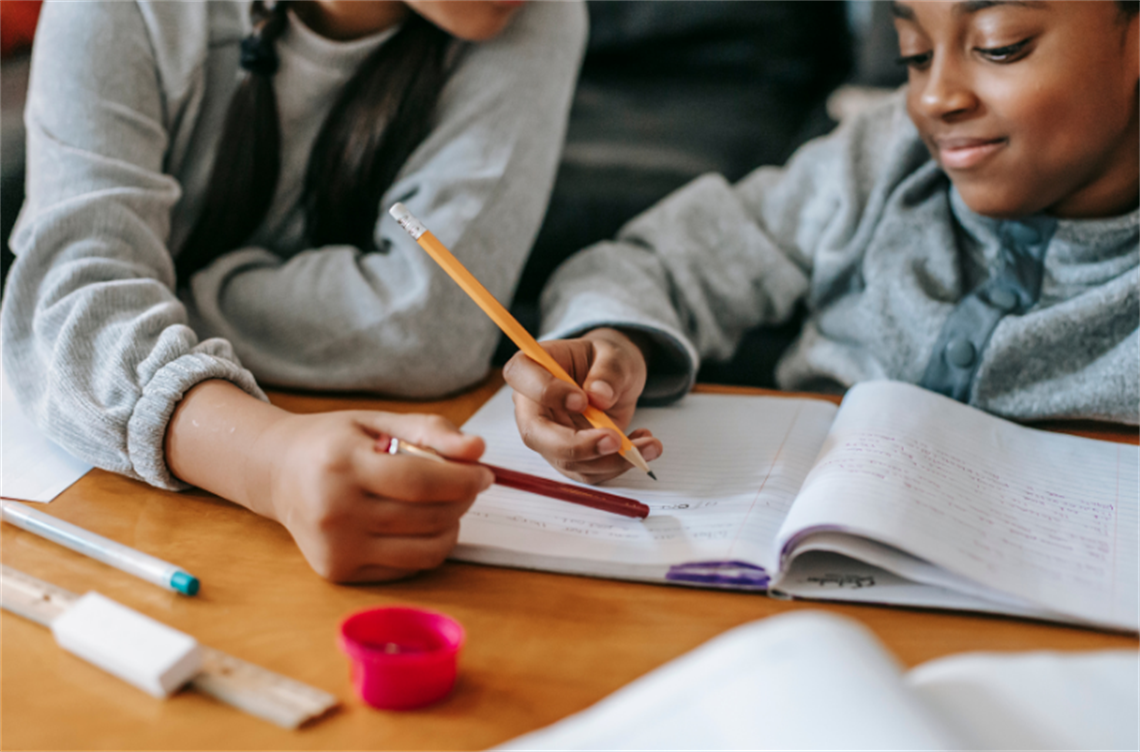 Two children collaborating on a homework assignment, writing in notebooks with pencils on a wooden table.