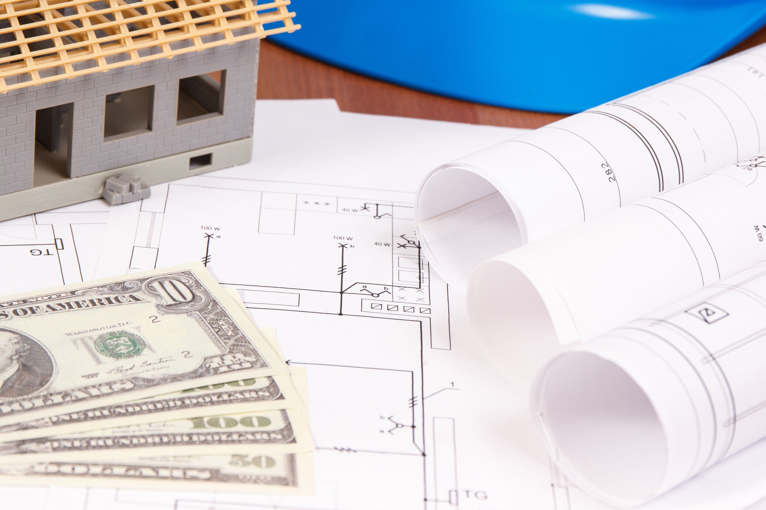 Home remodel blueprint, hard hat, and money signifying importance of cost with decision timing in remodels.
