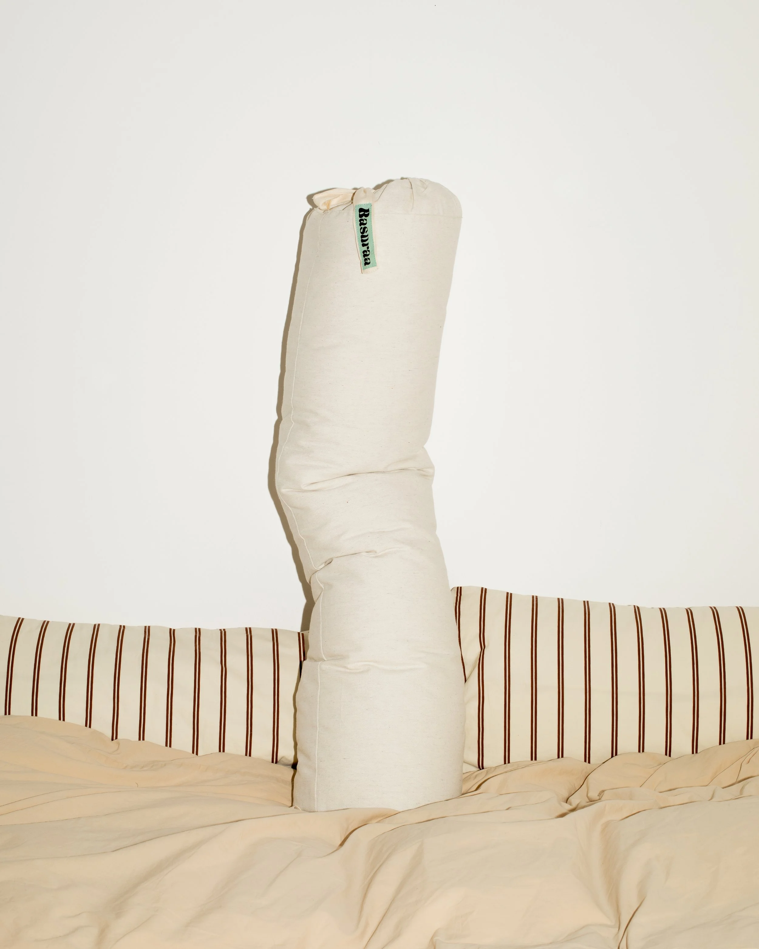 Ein gepolstertes großes Kisse, der wie eine große Zigarette aussieht, steht auf einem unordentlichen Bett mit gestreifter Kopfstütze. Der Gegenstand ist beige, hat ein türkisem Etikett mit schwarzer Schrift. Der Hintergrund ist eine weiße Wand.