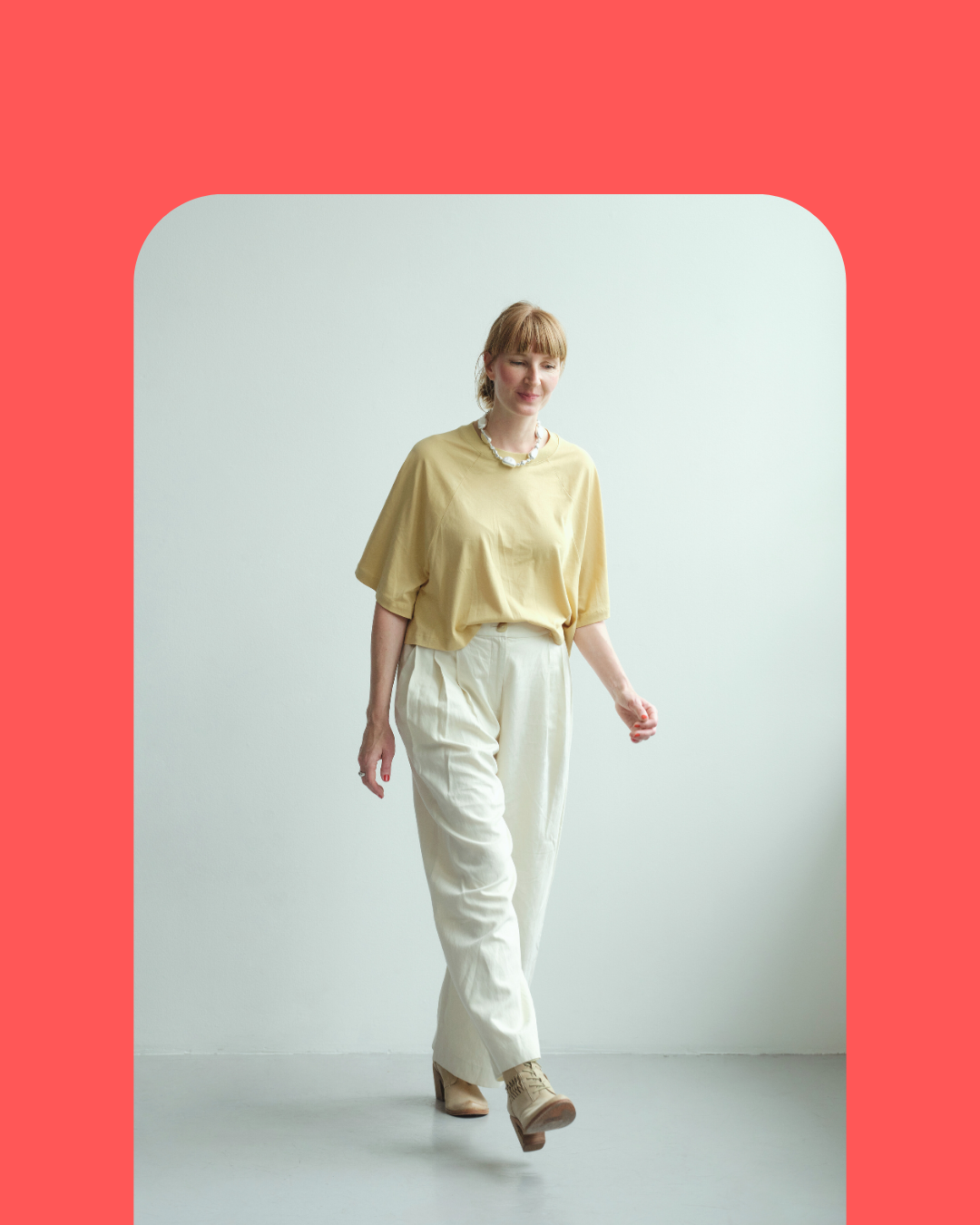 A woman walking inside, wearing a yellow top, cream-colored pants, and beige ankle boots, with a necklace, against a plain white background framed by a red border.