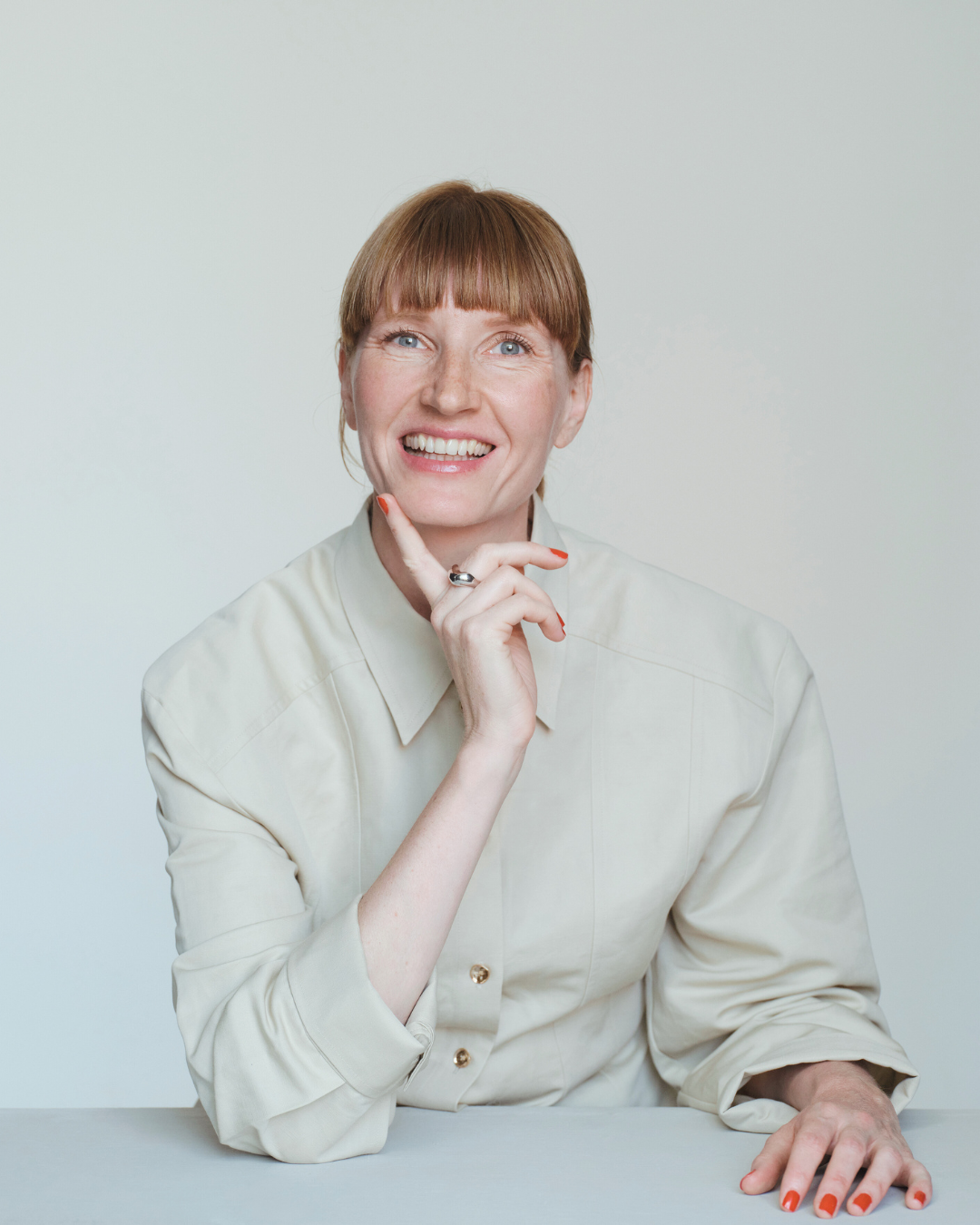 A woman with red hair and bangs smiling while resting her chin on her hand, wearing a cream-colored shirt, sitting at a table with a plain background.