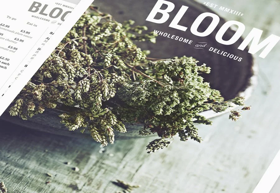A magazine with the title "BLOOM" and the subtitle "Wholesome and Delicious" placed on a light-colored wooden surface, with fresh green herbs, possibly thyme or oregano, spilling out of the magazine onto the surface.