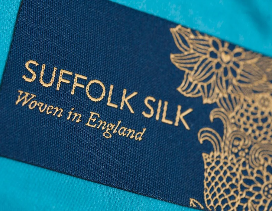 Close-up of a label on a piece of silk fabric, reading "Suffolk Silk" with decorative gold floral pattern, and text "Woven in England".