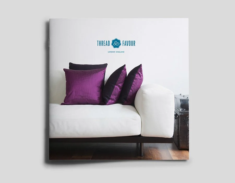 A white sofa with purple and black pillows, a sign that says "THREAD & FAVOUR" and "LONDON - ENGLAND" above it, and a metallic trunk or storage box beside it.