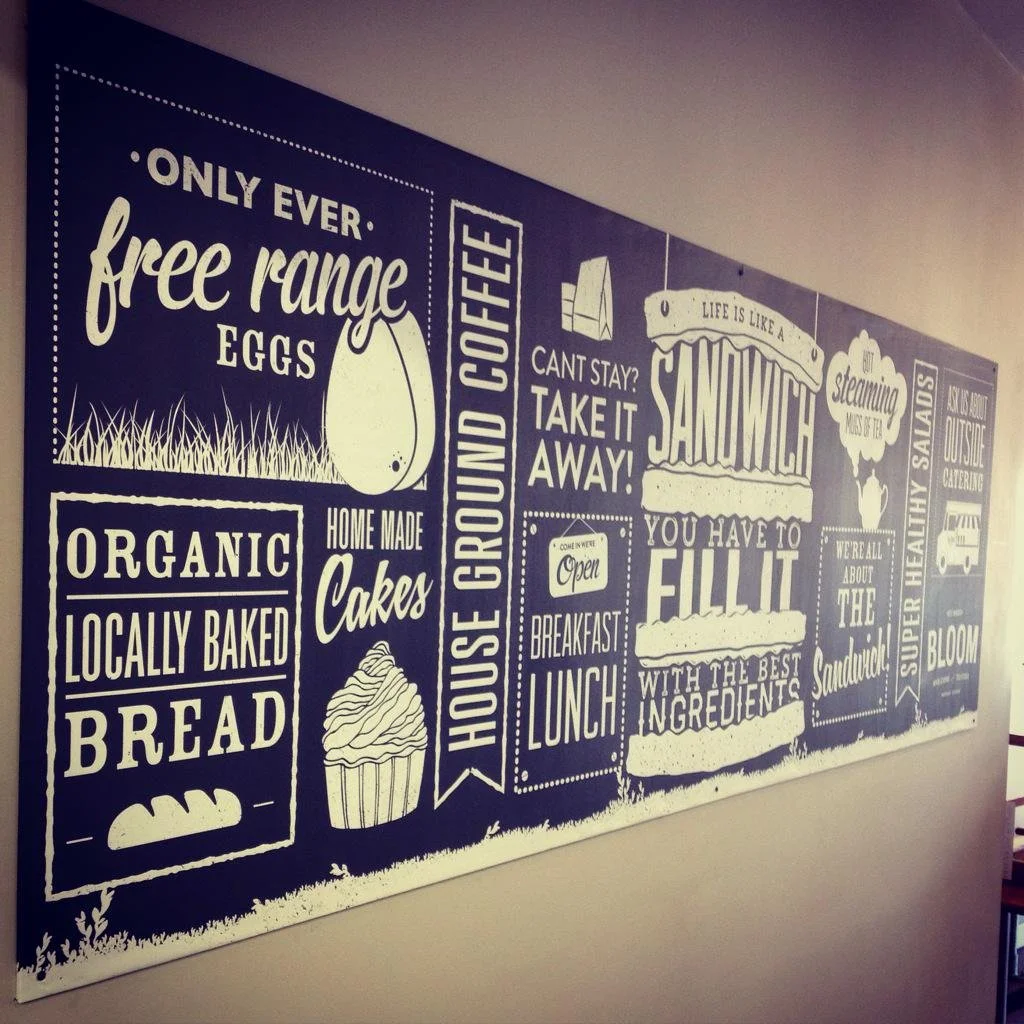 A wall chalkboard sign with various messages about a cafe, including references to free-range eggs, organic bread, home-made cakes, house ground coffee, and sandwich ingredients.