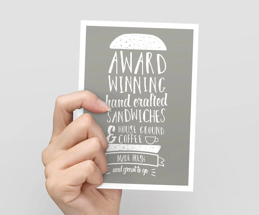 Hand holding a restaurant menu with handwritten text, advertising award-winning, handcrafted sandwiches, house ground coffee, and fresh ingredients.