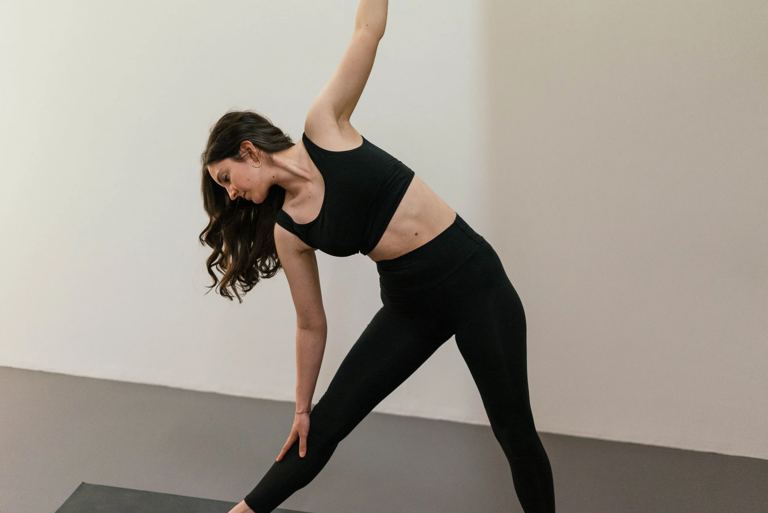 Eine Frau in schwarzer Sportkleidung macht eine Yoga- oder Dehnübung vor einer weissen Wand.