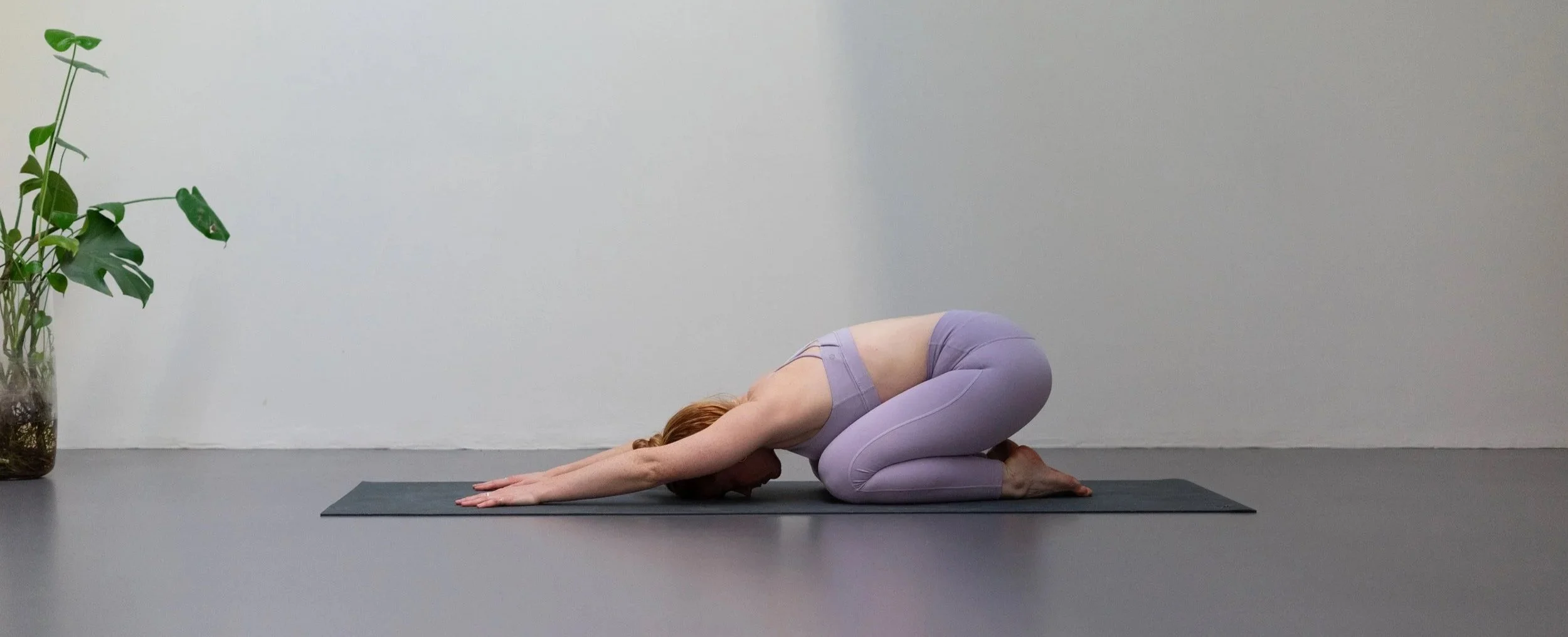 Frau in Childs Pose, auf einer Yogamatte in einem Raum mit grauer Wand und Topfpflanze auf der linken Seite.