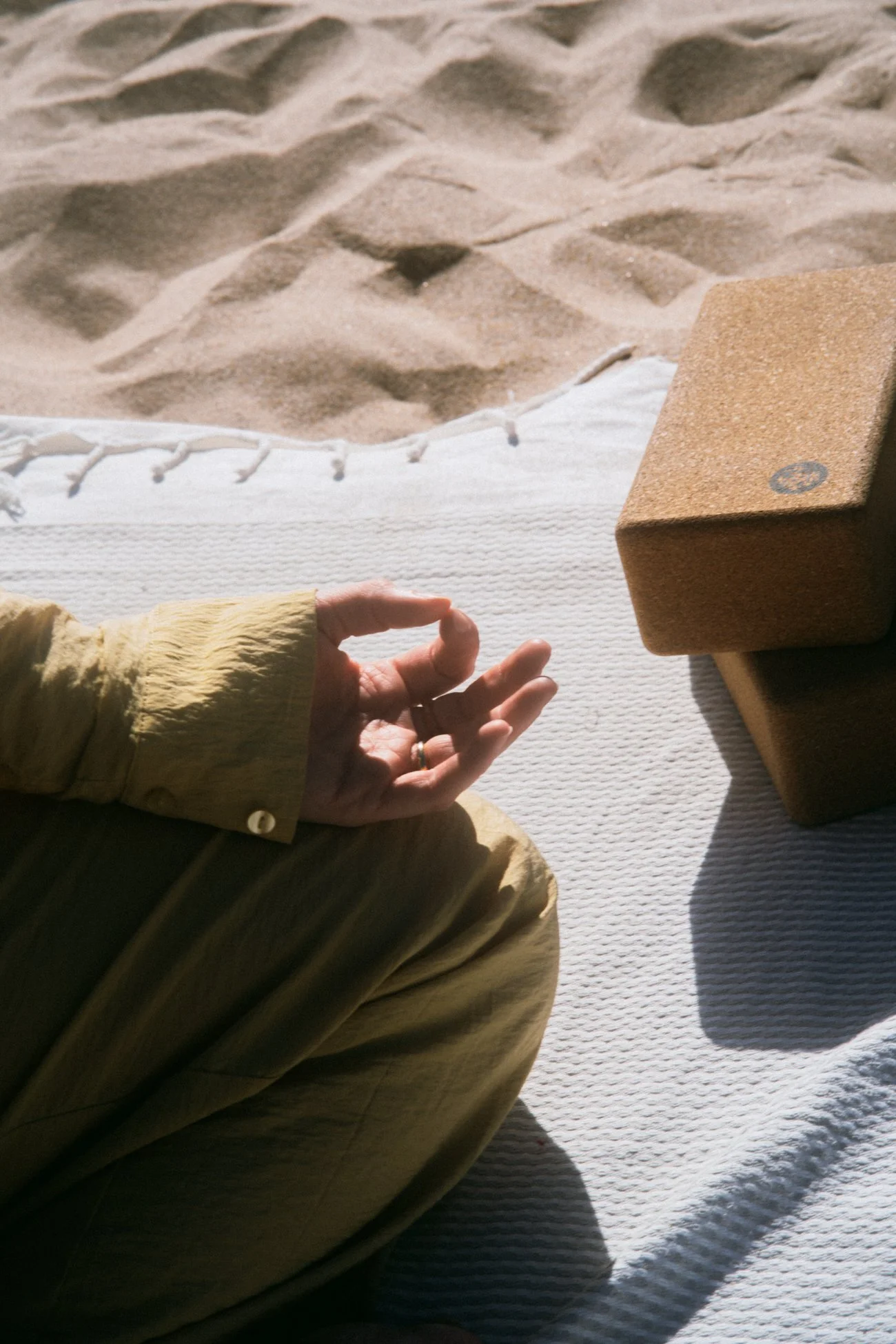Eine Person, die im Schneidersitz auf einem weißen Teppich sitzt, mit einem ausgestreckten Arm und einer Hand in der Meditation. Im Hintergrund befindet sich ein Sandhaufen neben einem Korkblock.