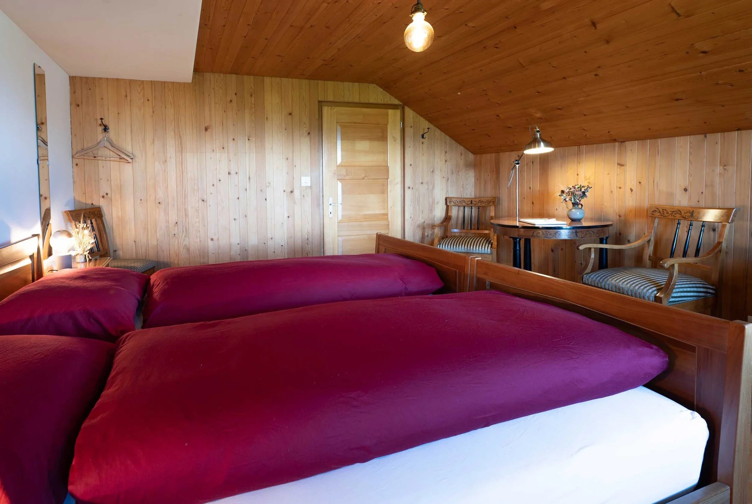 Ein Schlafzimmer mit Holzvertäfelungen an Wänden und Decke, einem Bett mit roter Bettdecke, einem kleinen Tisch mit einer Vase voller Blumen, mehreren Stühlen, einer steckdose und einer Leuchte.