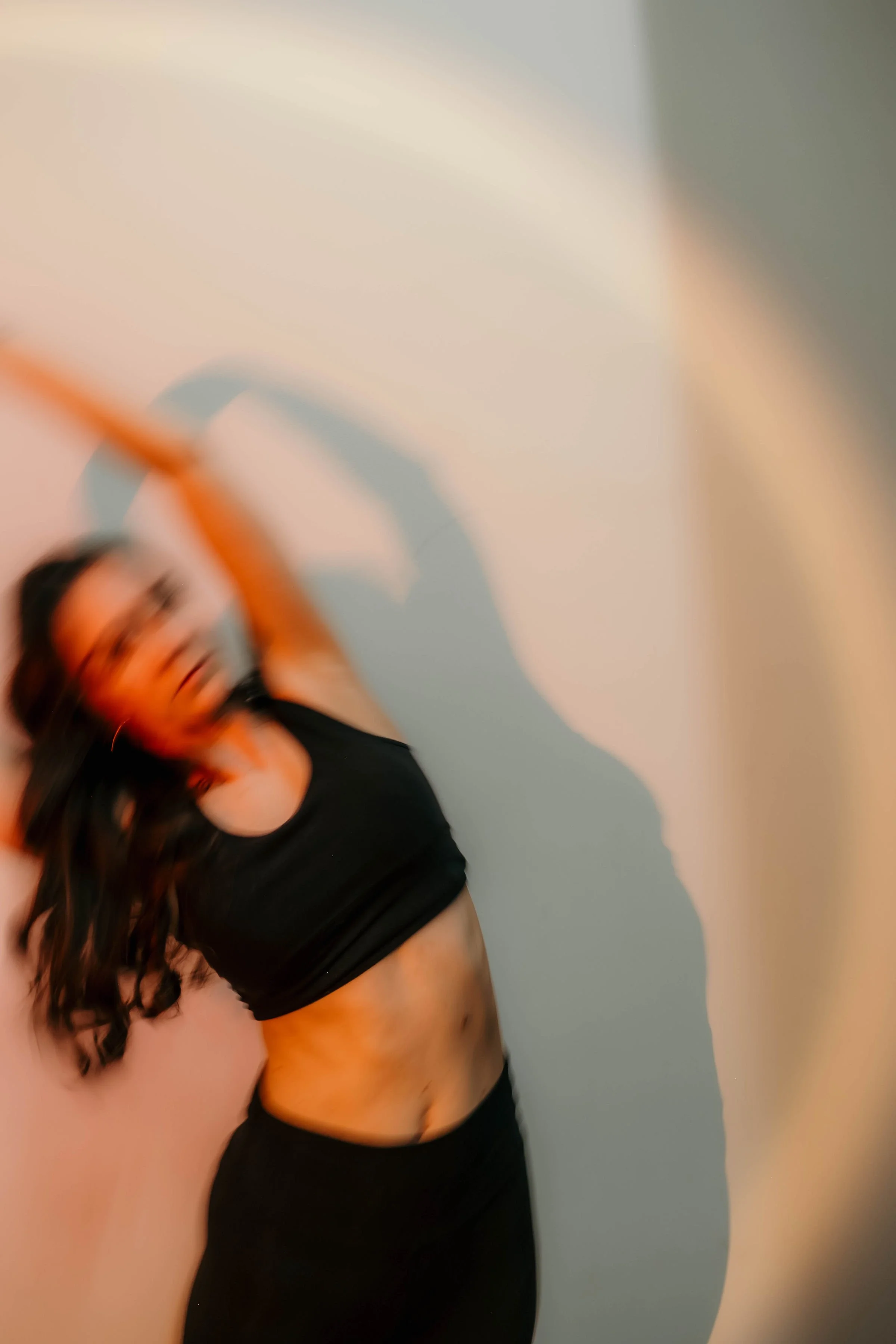 Eine Frau in schwarzem Sportoutfit, die ihre rechte Hand im Haar hat, steht vor einer zweifarbigen Wand, die oben beige und unten hellblau ist.