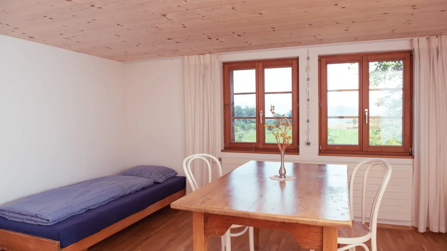 Ein einfaches Schlafzimmer mit einem Einzelbett, einem holztisch, zwei weissen Stühlen, einem Fenster mit Blick auf grüne Landschaft, weißen Vorhängen und Holzboden.