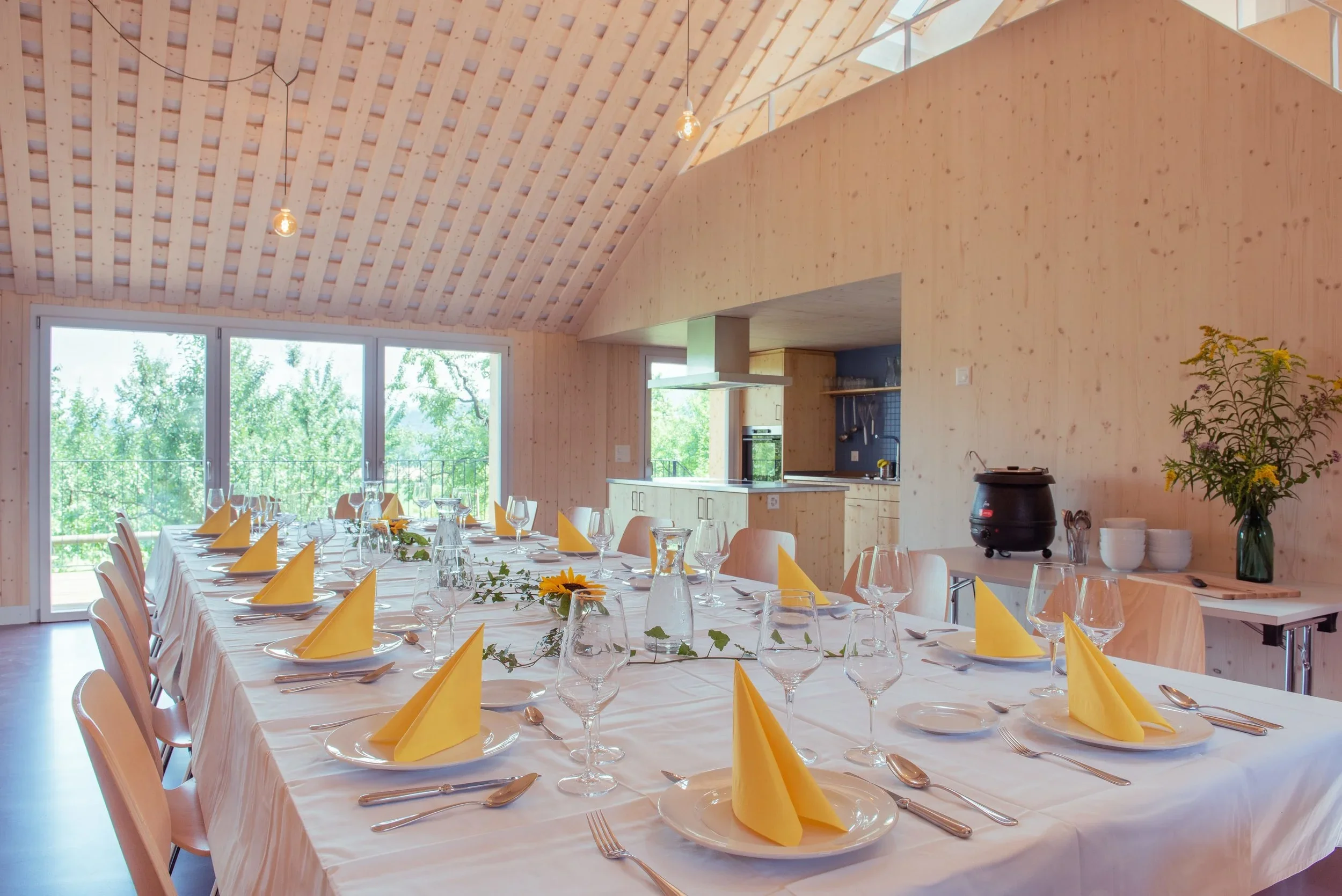 Ein festlich gedeckter Esstisch mit weißen Tischtüchern, gelben Stoffservietten und Gläsern in einem modernen, lichtdurchfluteten Raum mit Holzinterieur und grossen Fenstern.