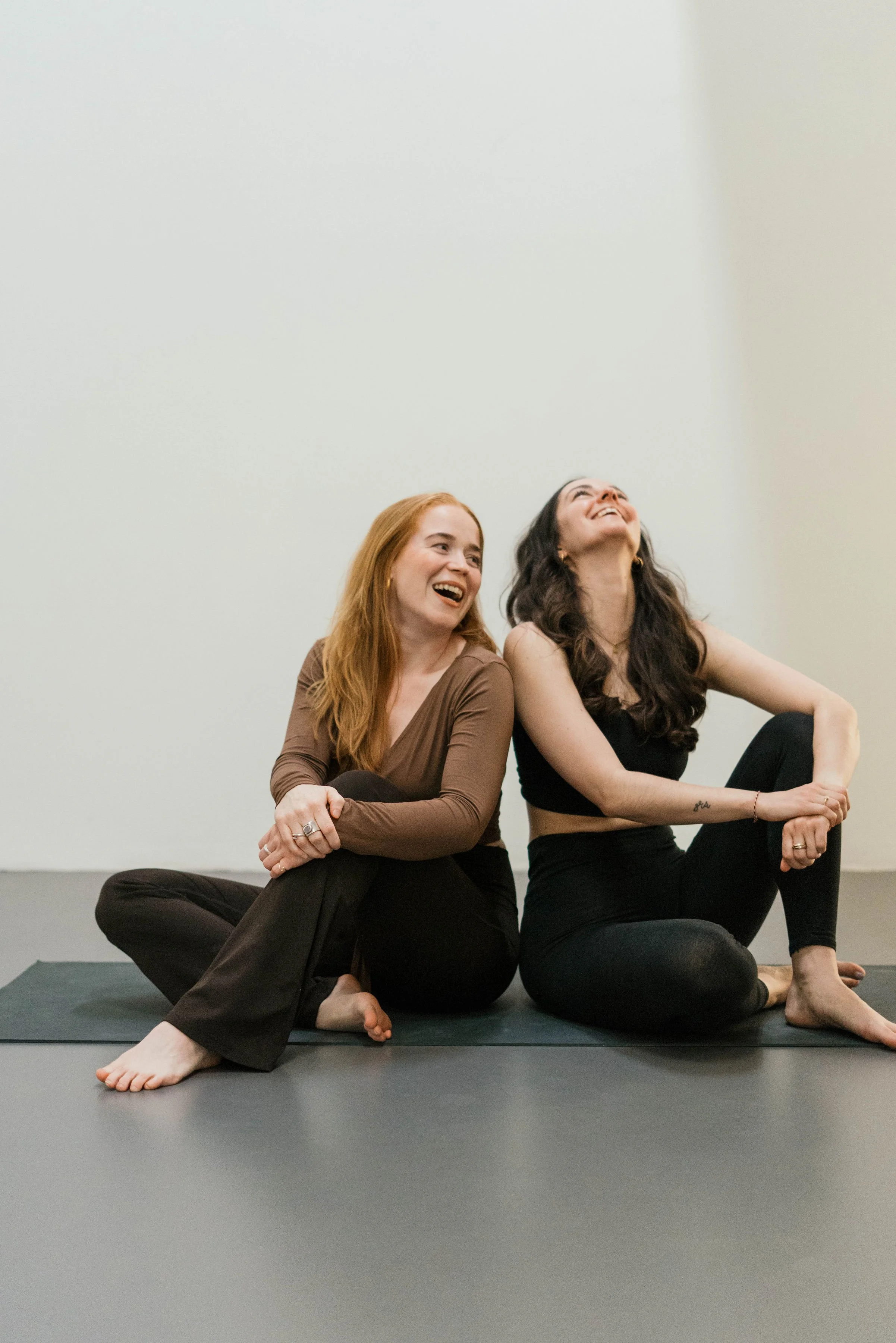 Zwei Frauen sitzen entspannt auf dem Boden und lachen gemeinsam, während sie auf einer Yogamatte in einem hellen Raum sitzen.