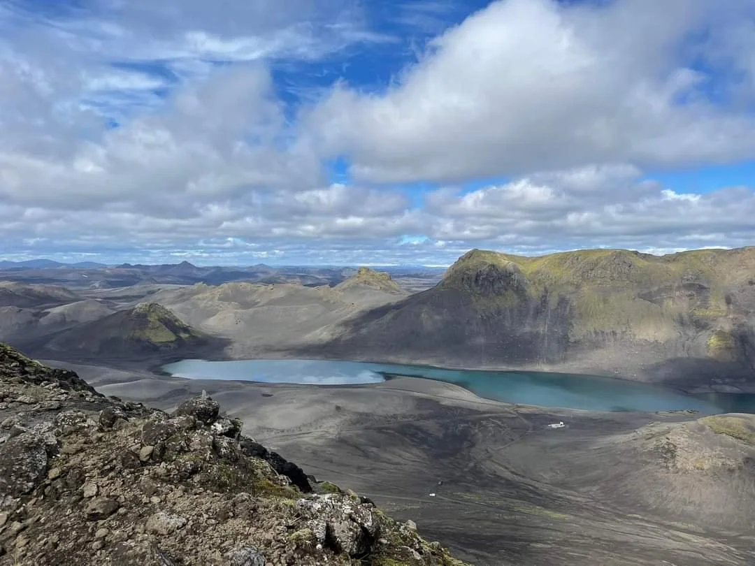 A volcanic crater with two lakes, surrounded by rugged mountains under a partly cloudy sky in a remote, barren landscape.