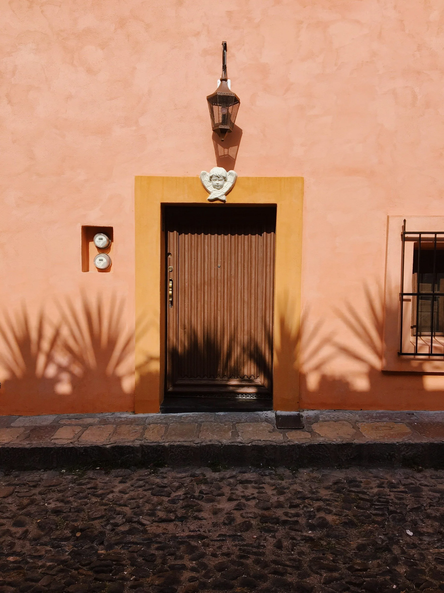 A wooden door set in a peach-colored wall with a yellow frame, with an angel face decoration above it, and a lantern hanging above that. Shadows of palm leaves are cast on the wall to the sides, and there are two outlets on the left side of the wall.