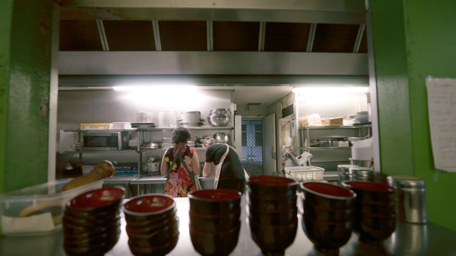 View through a kitchen window showing two people working in a commercial kitchen, with stacks of red bowls on the foreground counter.
