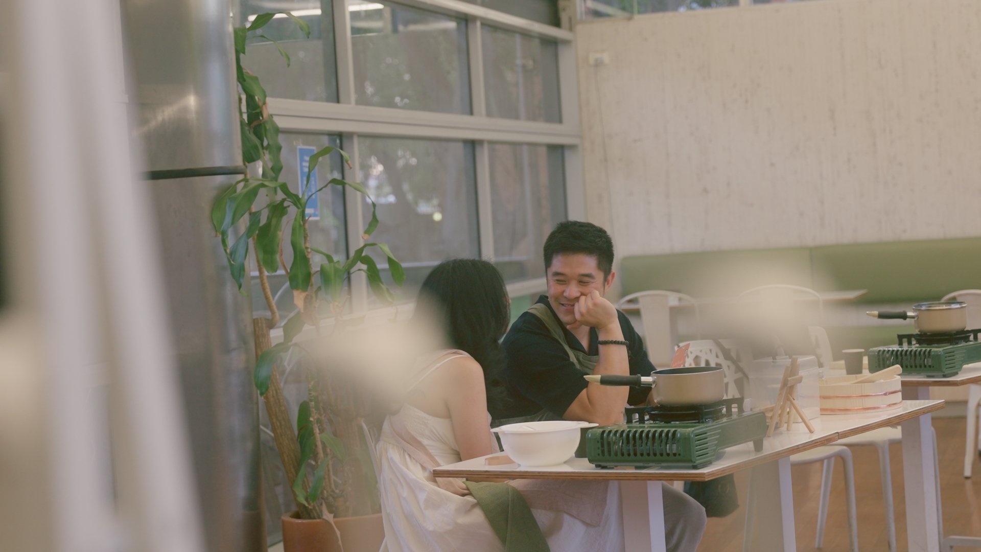 A man and woman sitting at a table in a cafe, smiling and talking to each other with a window and plants in the background.