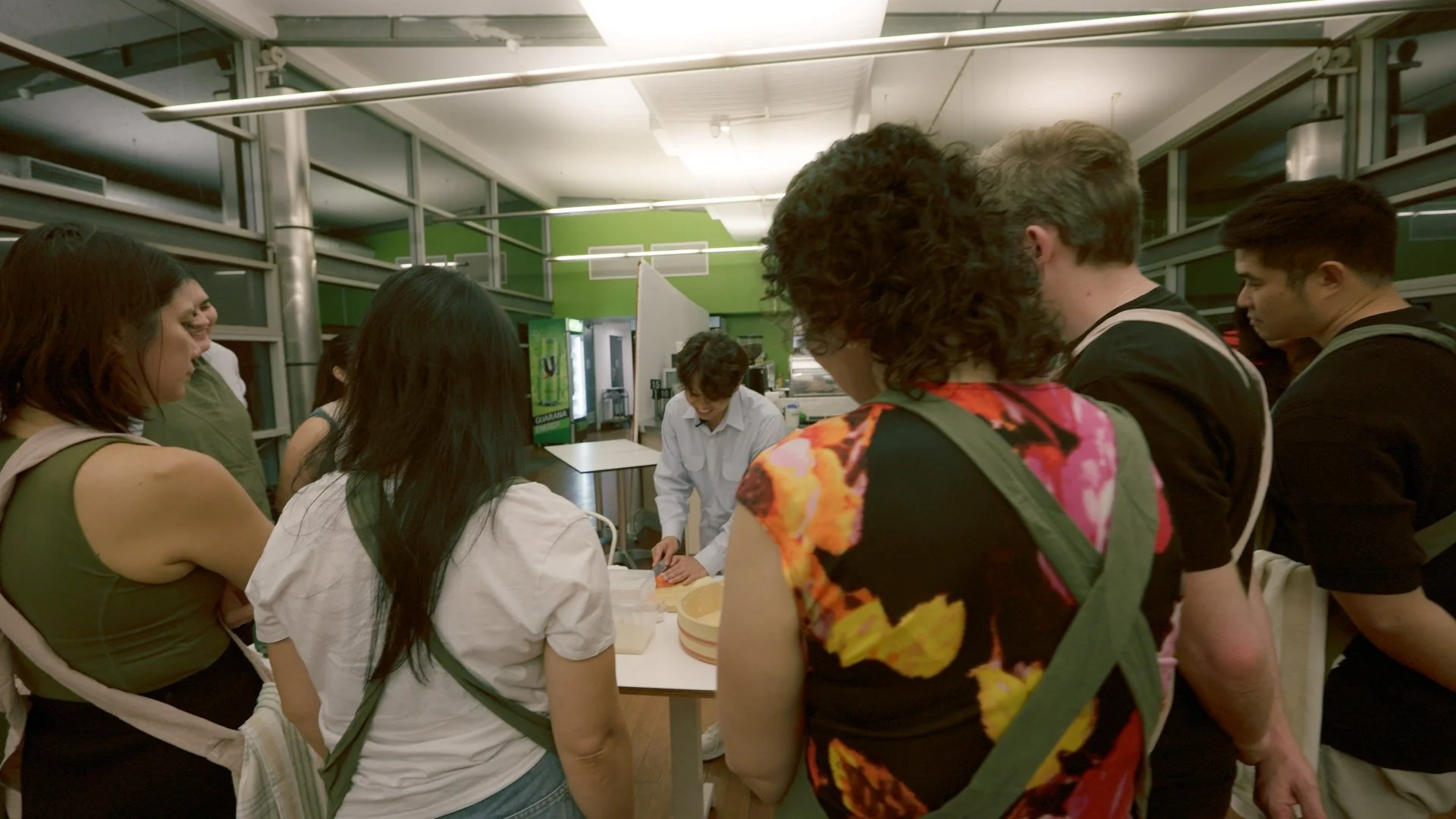 Group of people gathered around a table in a kitchen or food prep area, with a person slicing or preparing food, surrounded by onlookers.