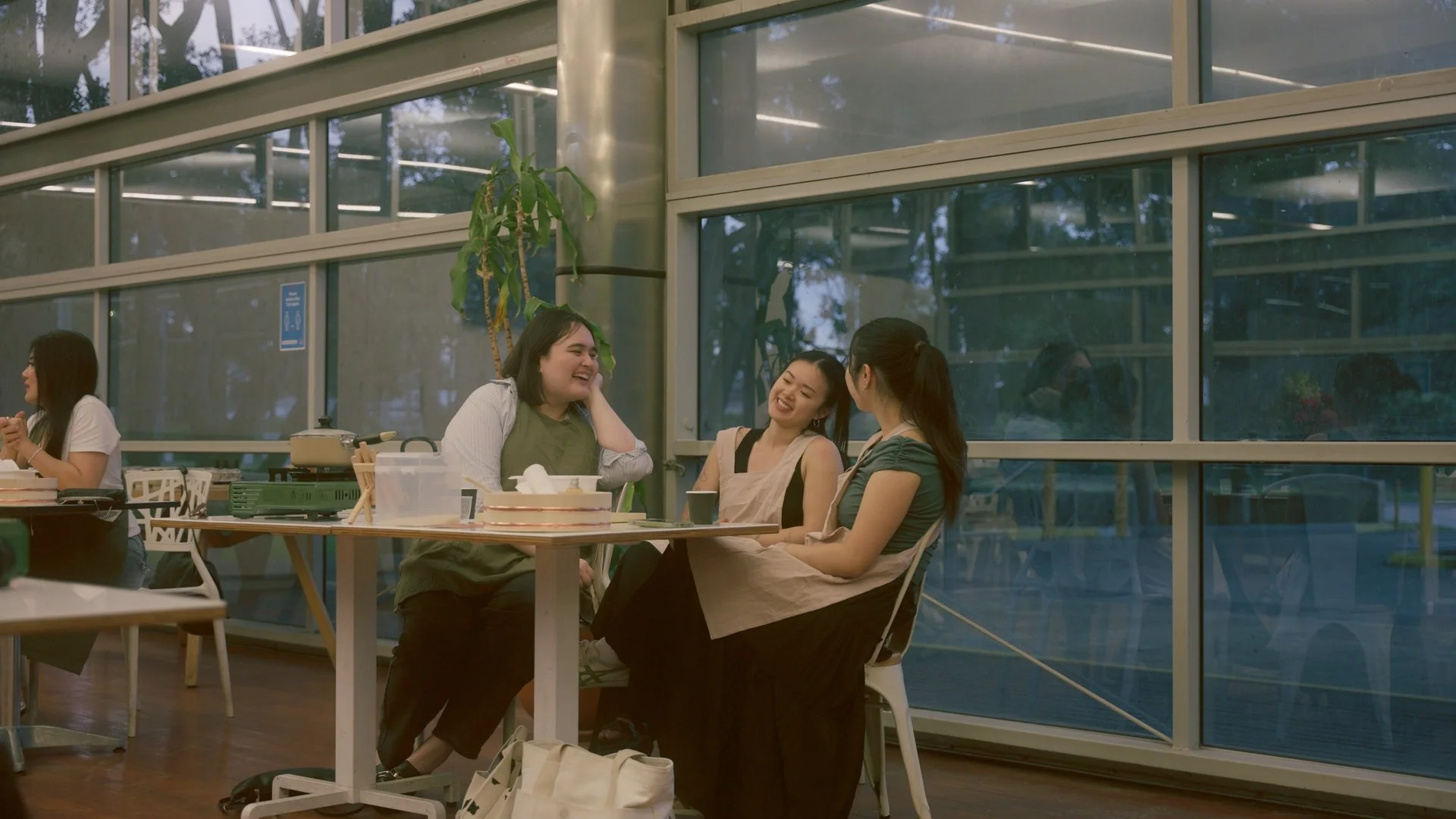 Three women sitting at a table inside a greenhouse-like structure, engaged in conversation and laughing, with a woman in the background sitting alone.
