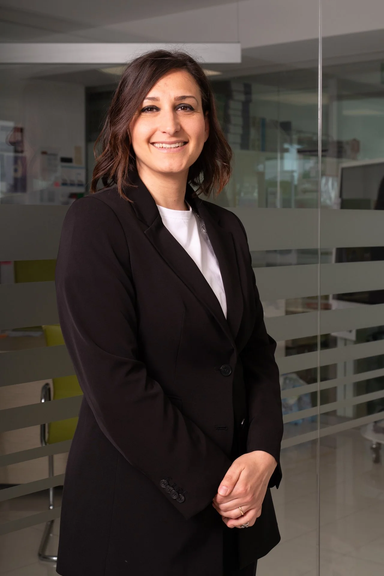 Una donna sorridente con capelli castani riccioli, indossa un abito da lavoro nero, si trova in un ufficio moderno con pareti di vetro e scrivanie.