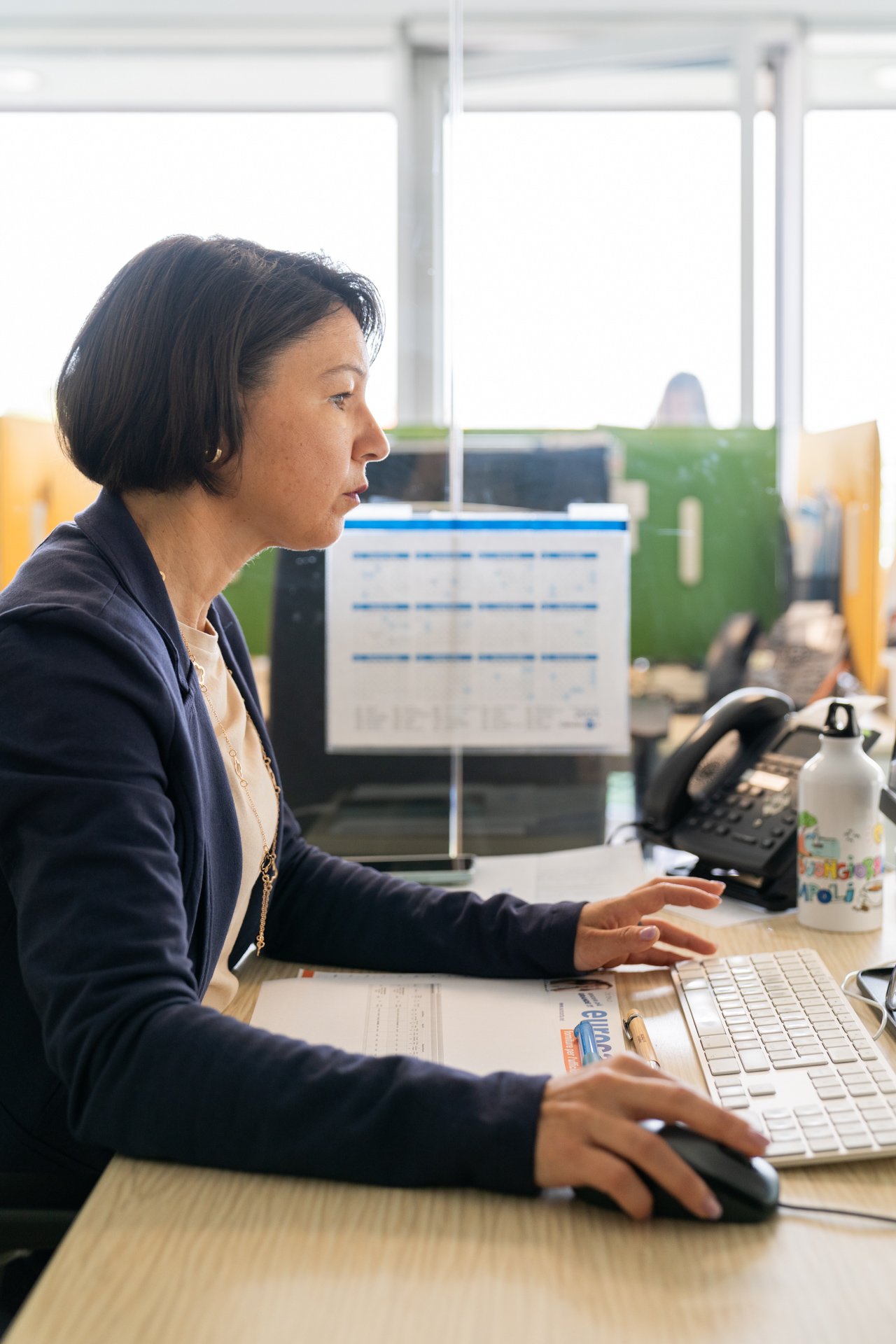 Una donna che lavora in un ufficio, seduta davanti a una tastiera e un mouse, con telefono, monocchio, e vari oggetti da ufficio sul tavolo, con divisioni in vetro che separano le postazioni.