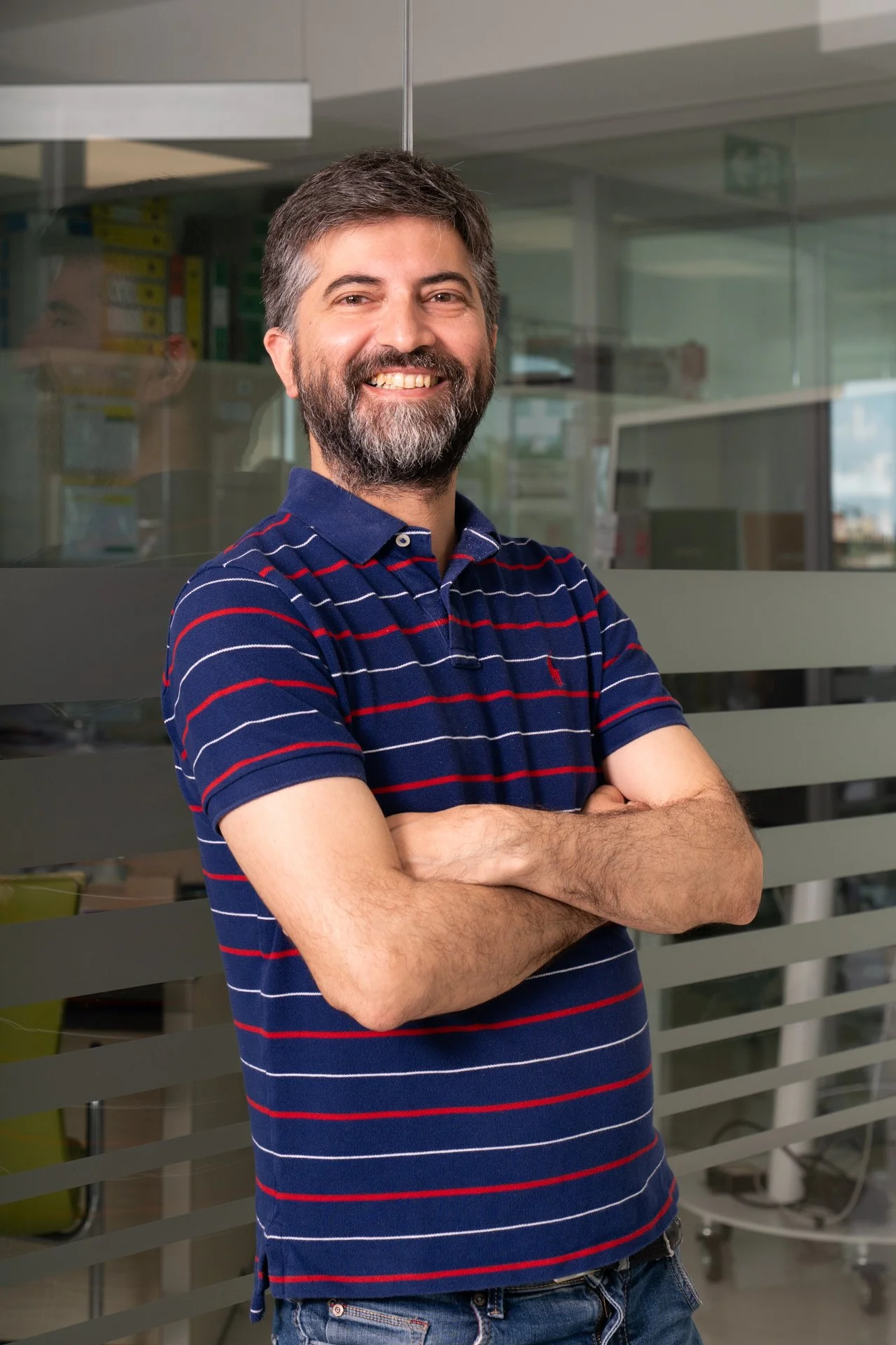 Uomo sorridente con barba e capelli corti, indossa una polo blu con righe rosse e bianche, in piedi con le braccia incrociate in un ambiente di ufficio moderno.