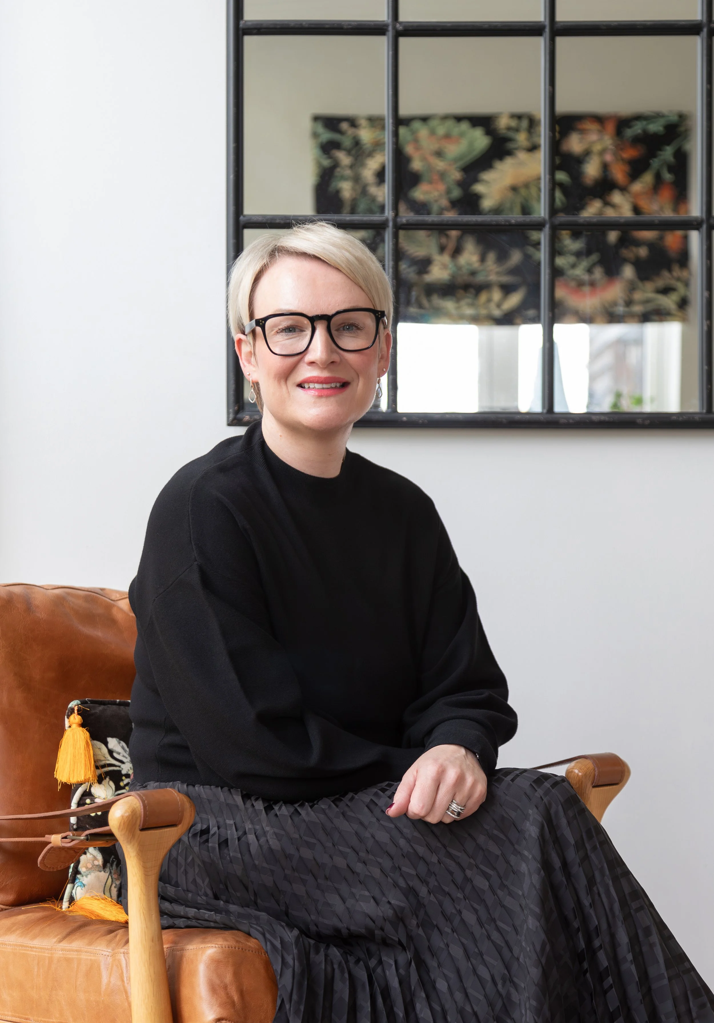 Alex Akrimi smiling while seated in a tan leather armchair, wearing a black top and dark pleated skirt