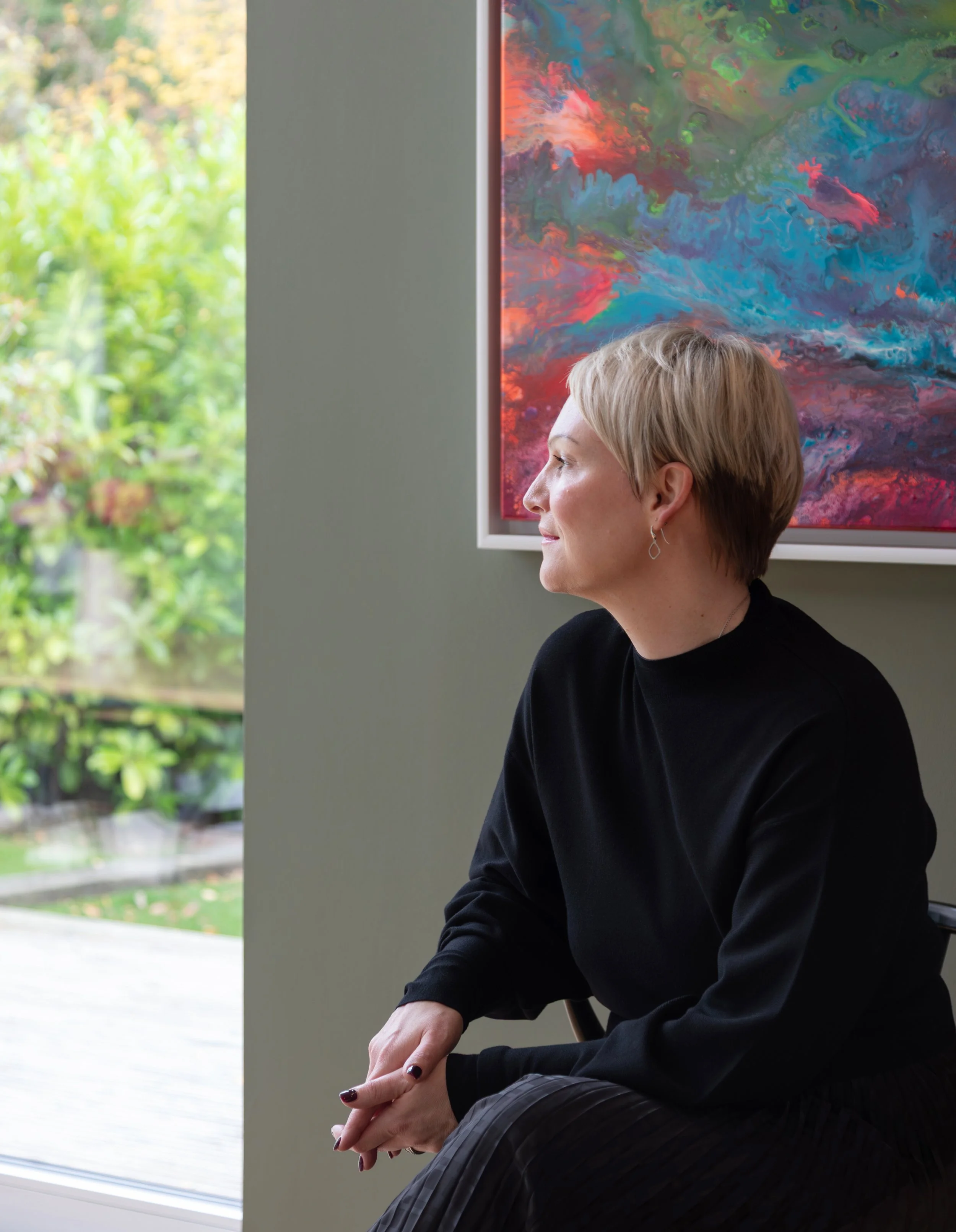 Alex Akrimi seated in profile, looking out of the window, wearing a black top and dark skirt beside colourful artwork