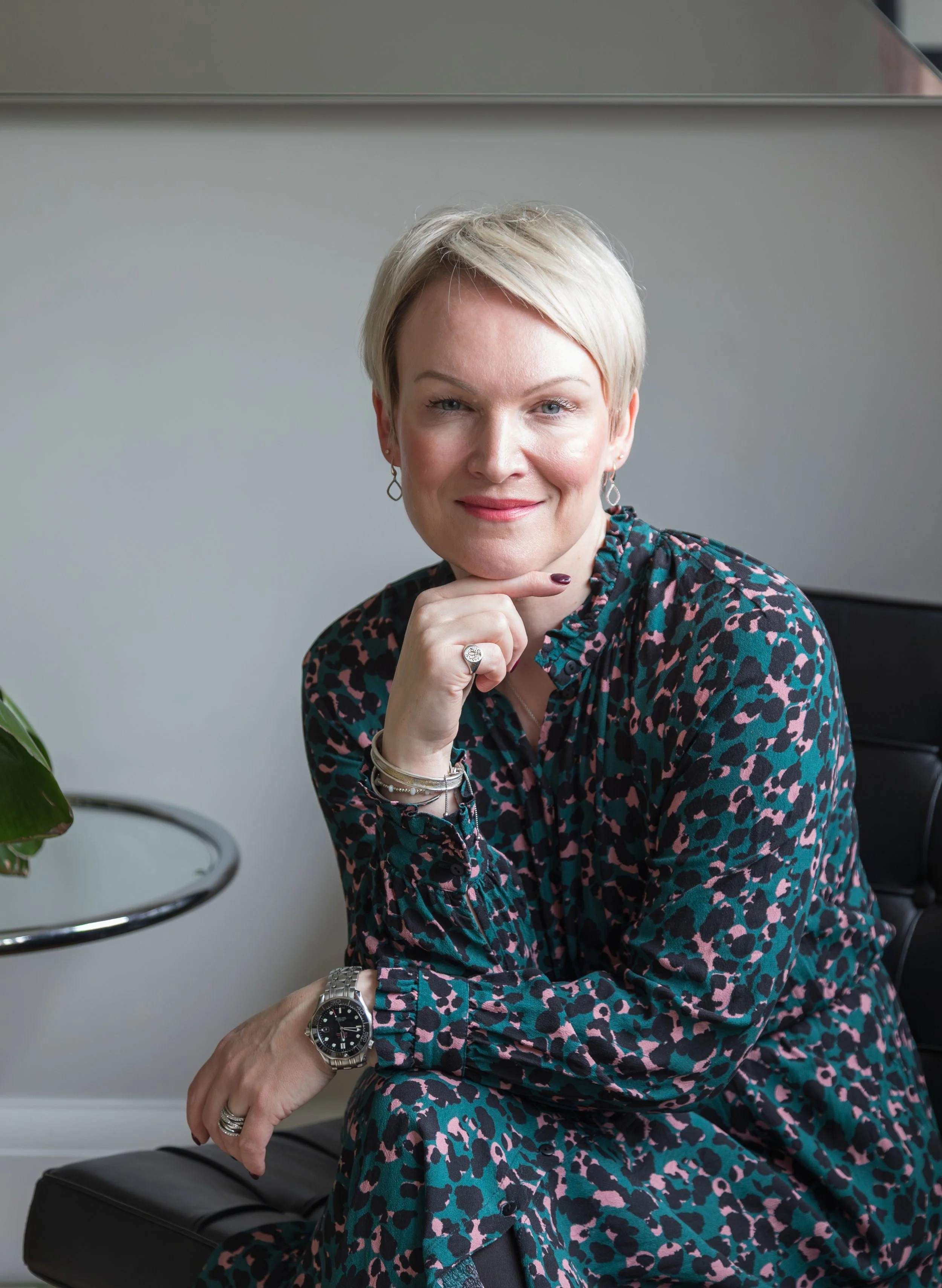 Alex Akrimi seated in a black chair, smiling with hand on chin, wearing a green and pink patterned dress