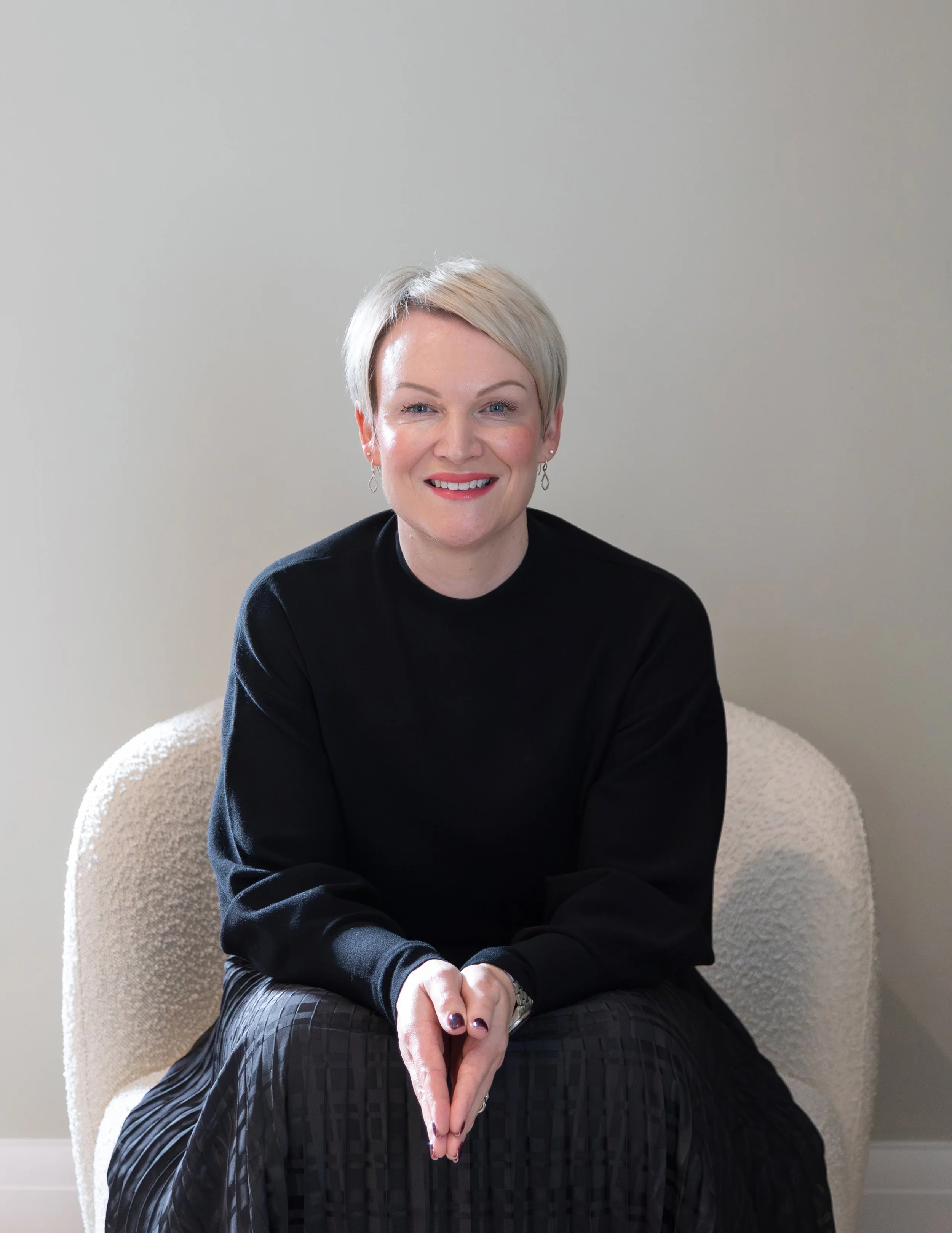 Alex Akrimi seated and smiling in a black top against a soft neutral background