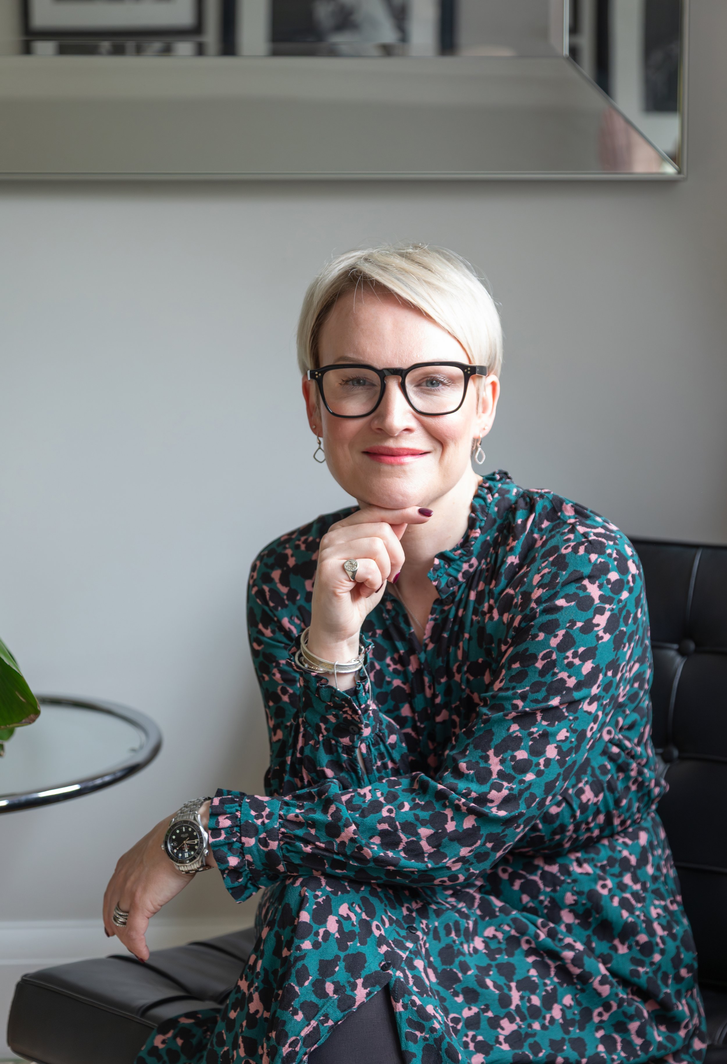Alex Akrimi seated and smiling in a green patterned dress, wearing glasses and resting hand on chin
