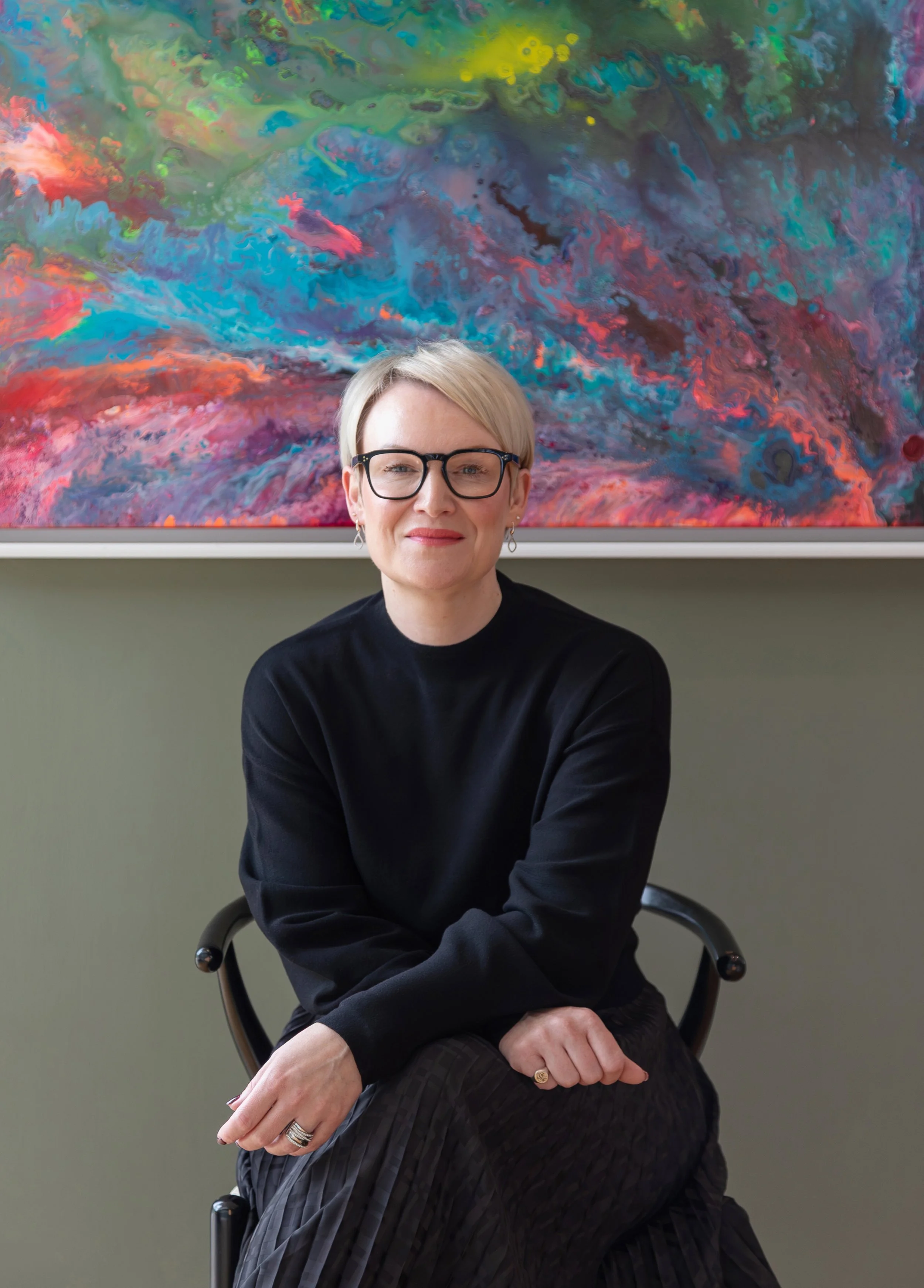 Alex Akrimi seated in front of a vibrant abstract painting, wearing glasses and a black jumper, smiling gently.