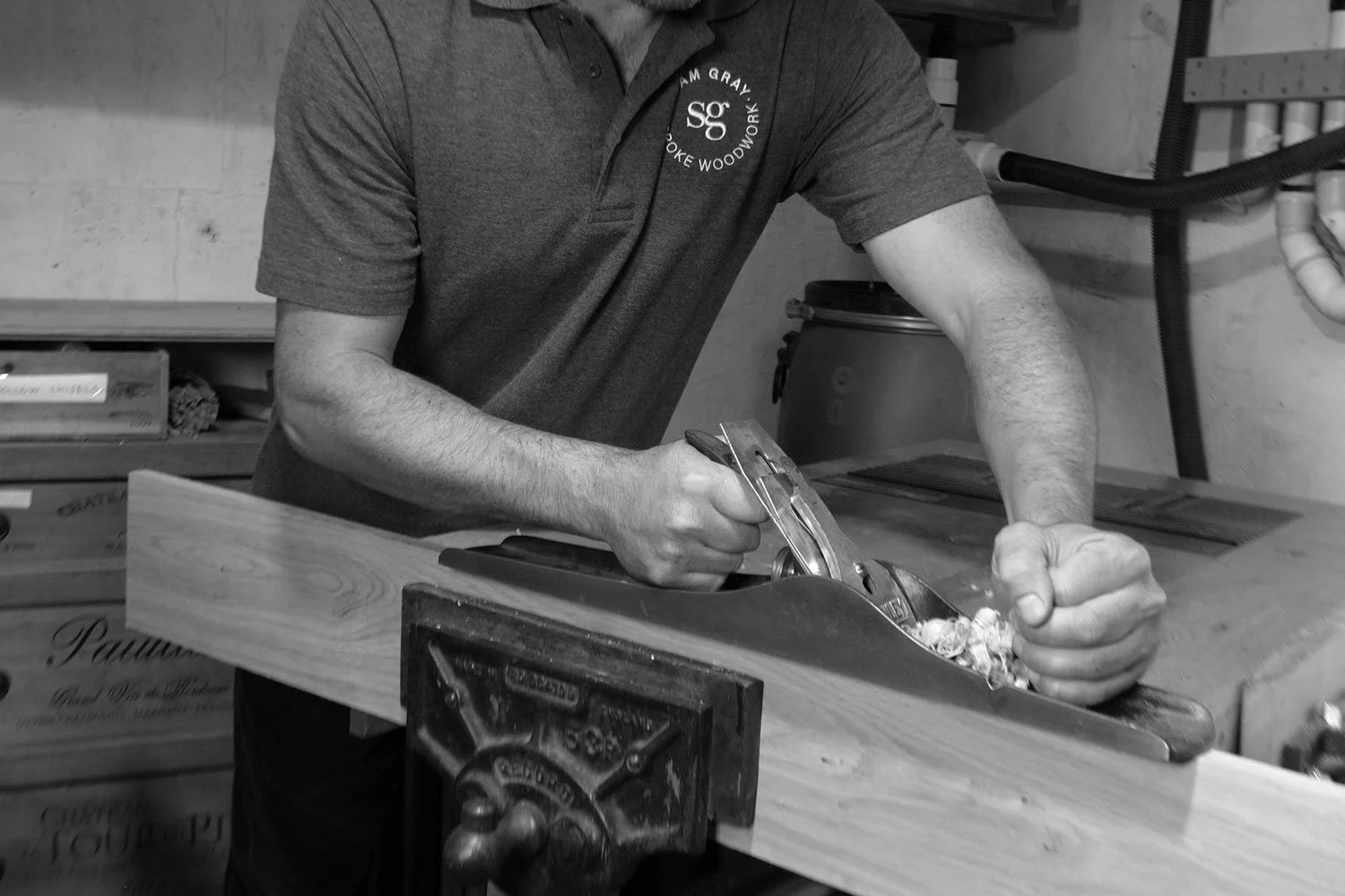 A black and white photo of a man focused on shaping a piece of wood with tools in a workshop setting.