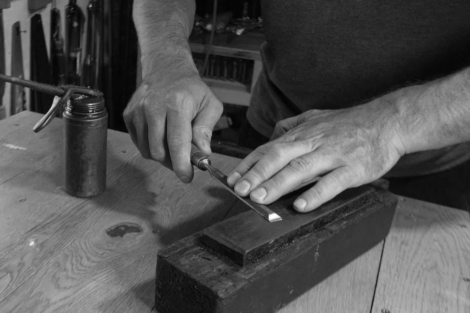 A black and white photo of a man focused on shaping a piece of wood with tools in a workshop setting.
