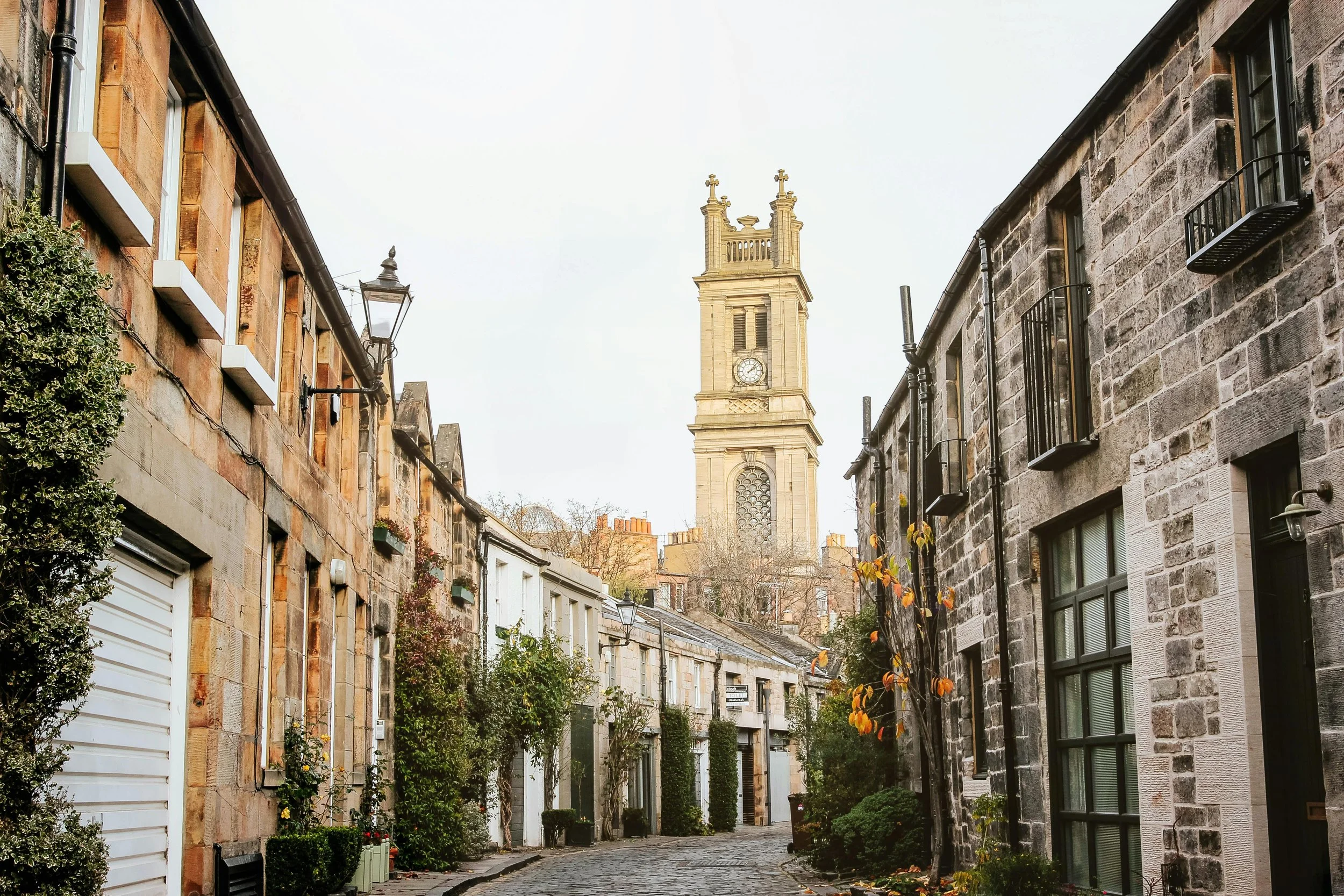 A cobblestone street lined with historic stone buildings and greenery, leading to a tall clock tower in the background.