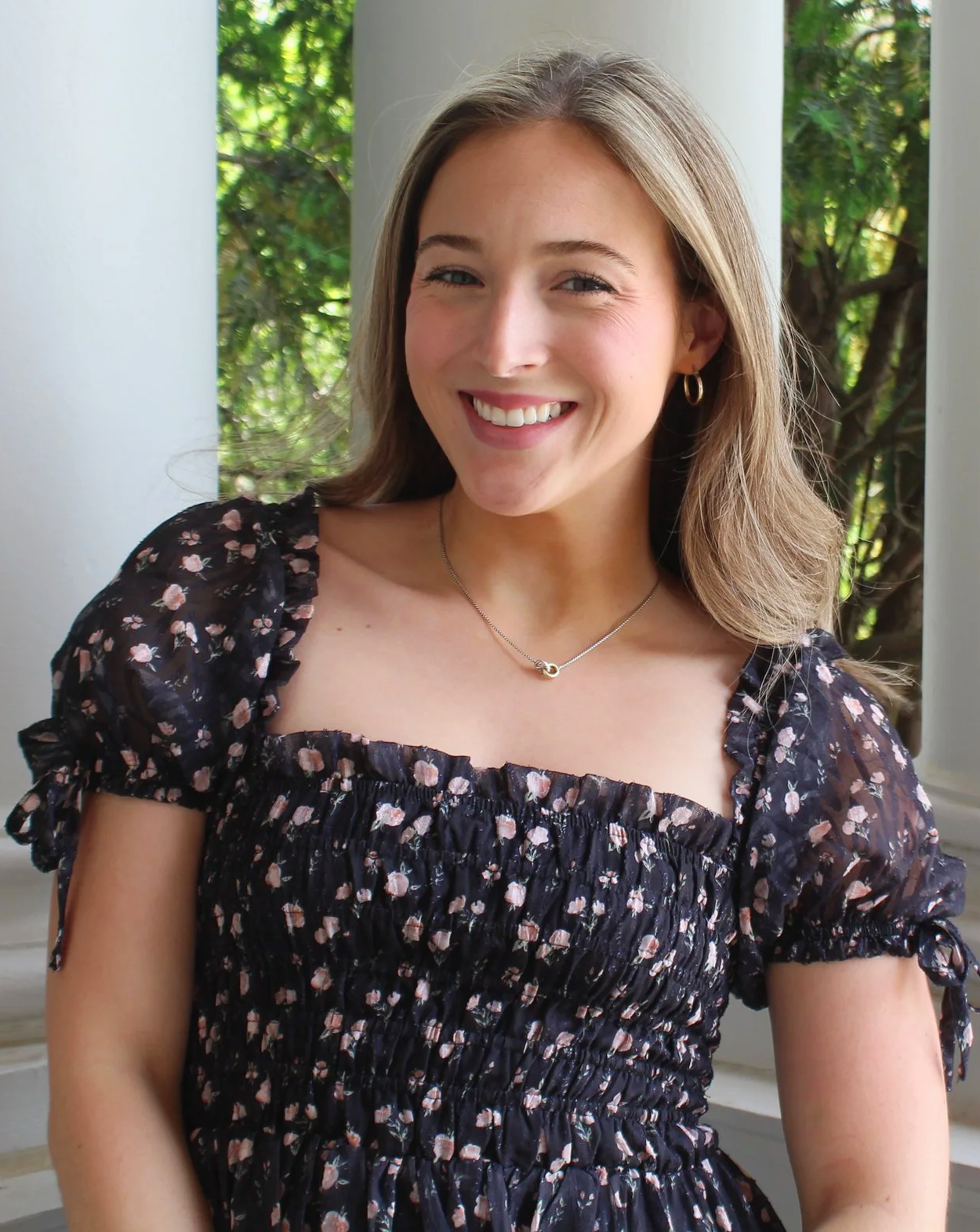 Sara Moyers smiling, wearing a floral black dress with puffy sleeves, gold hoop earrings, and a necklace, standing outdoors with greenery in the background.