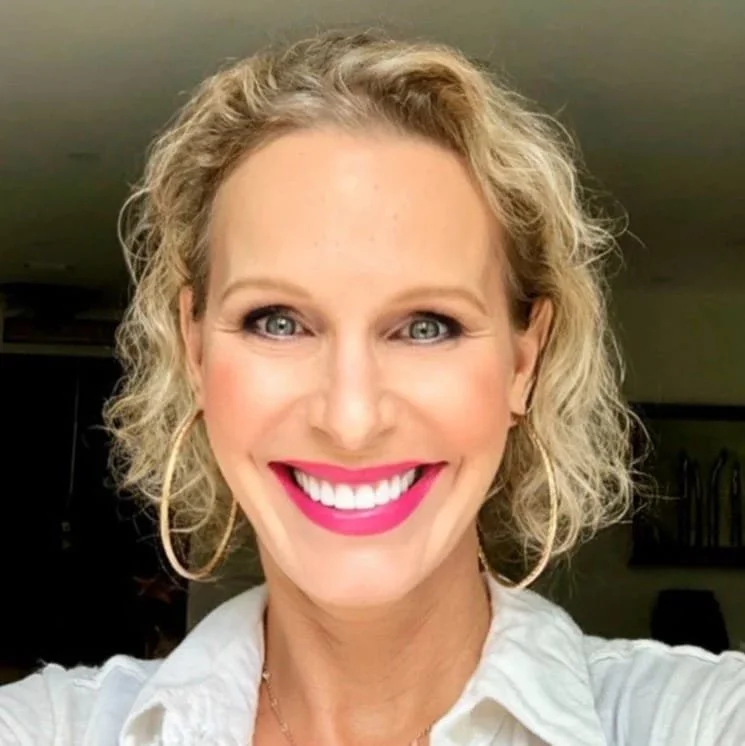 Danna Stillman's headshot with blonde curly hair, wearing pink lipstick, hoop earrings, and a white blouse, in an indoor setting.
