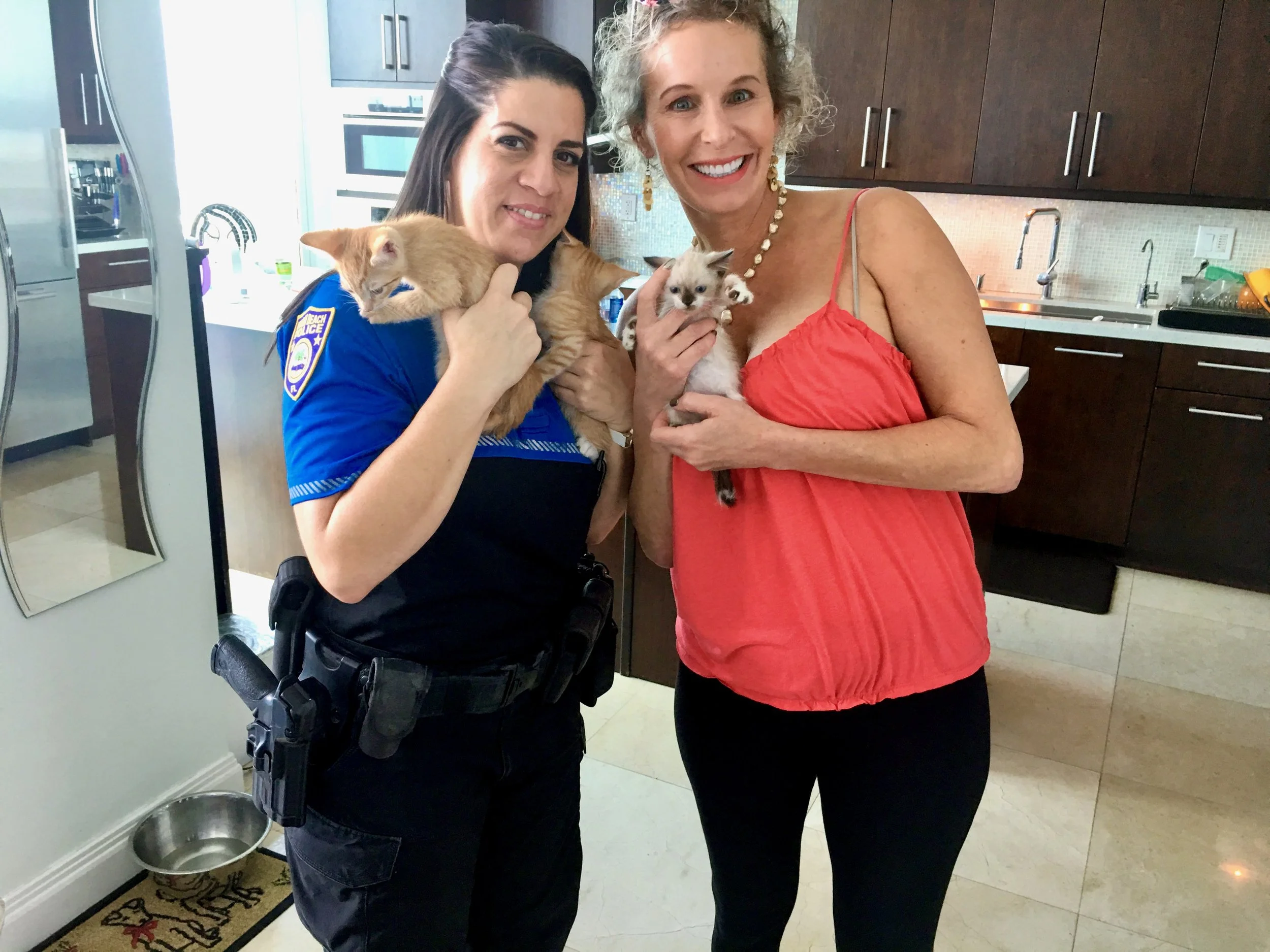 A woman in a police uniform holding an orange tabby kitten, standing next to another woman in a red sleeveless top holding a white and gray kitten, in a kitchen with dark wooden cabinets and a stainless steel sink.